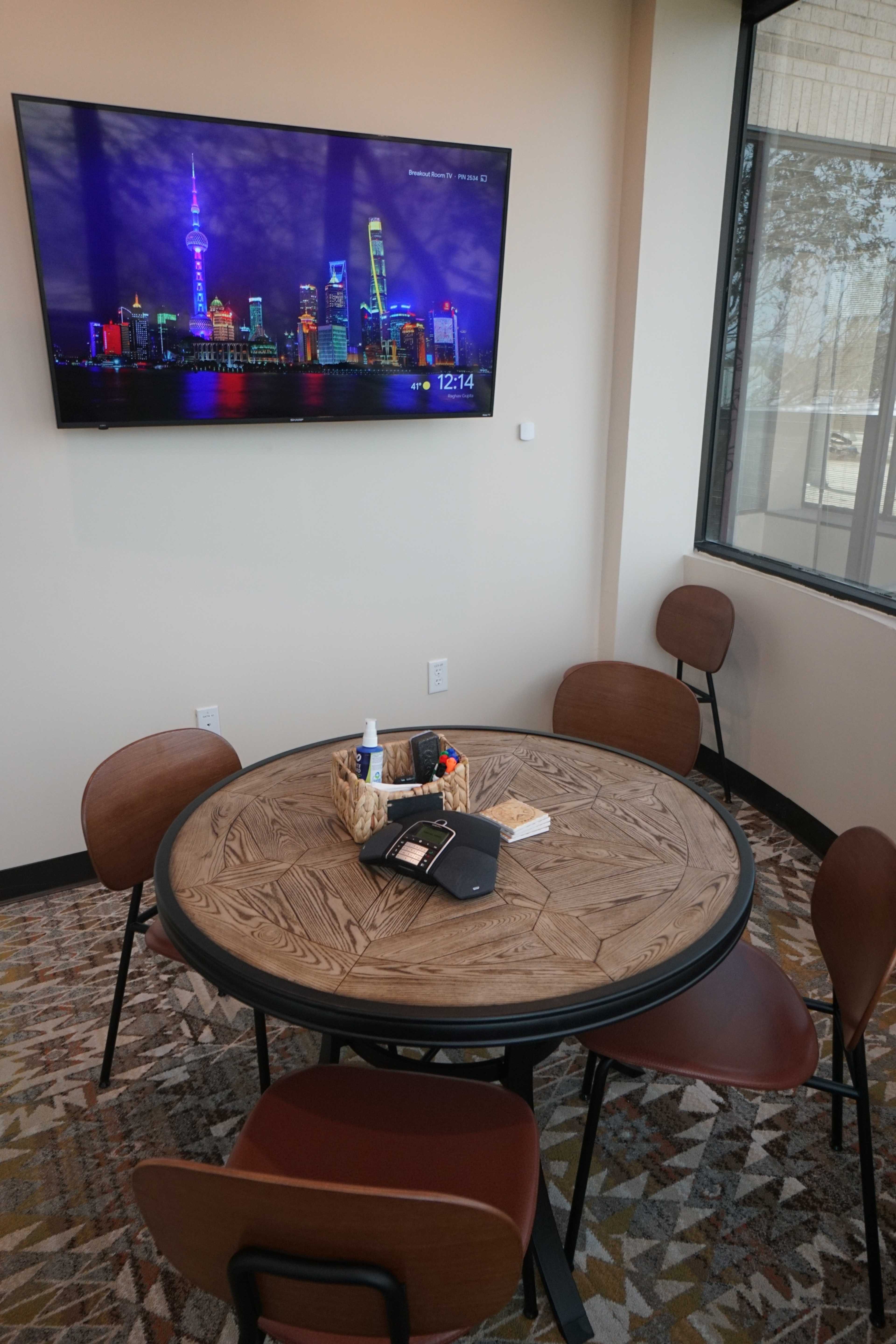 A round wooden table with four chairs is set in a room featuring a large TV displaying a city skyline at night.
