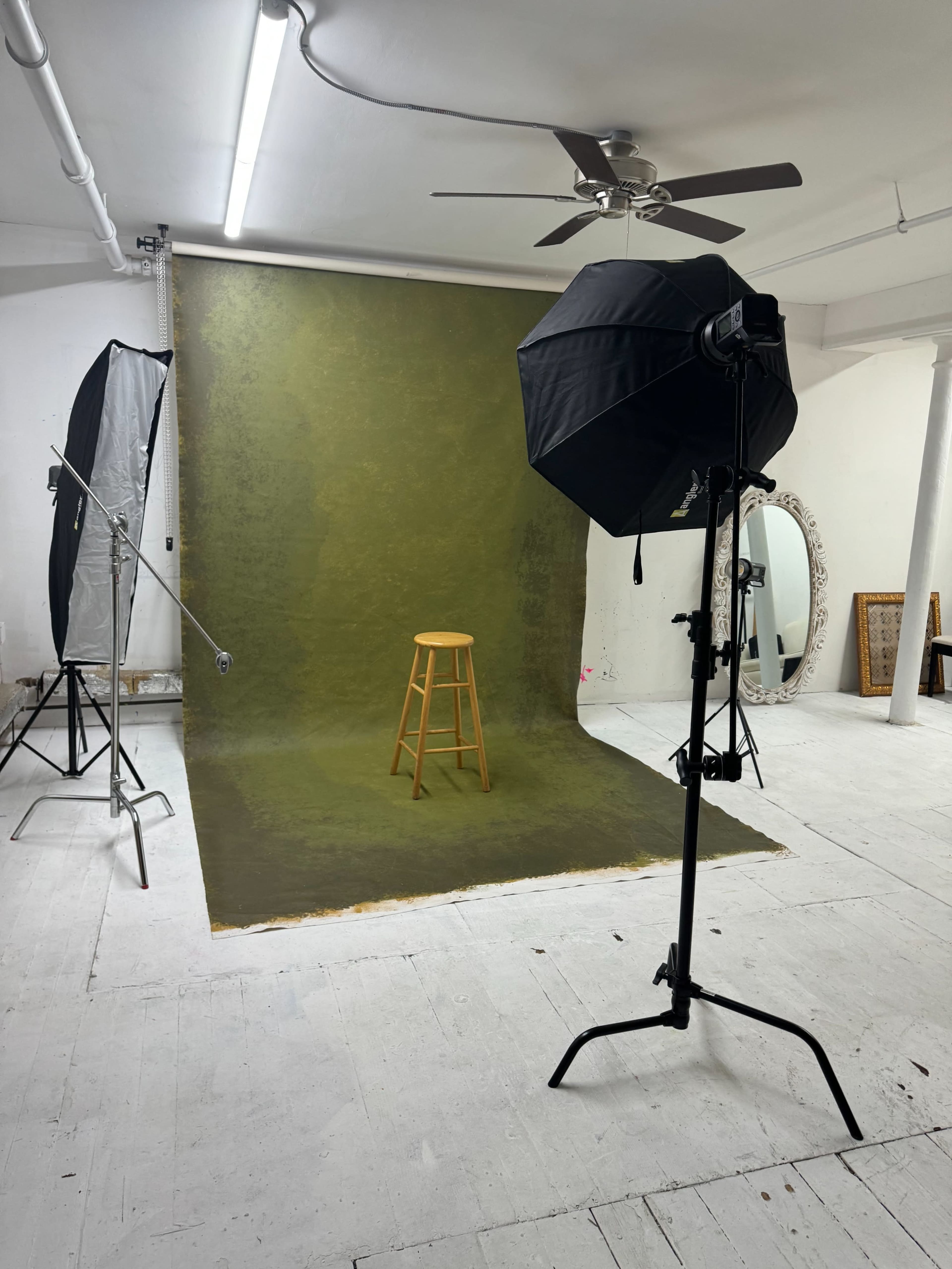A photography studio features an empty wooden stool in front of a green backdrop, with lighting equipment set up nearby.