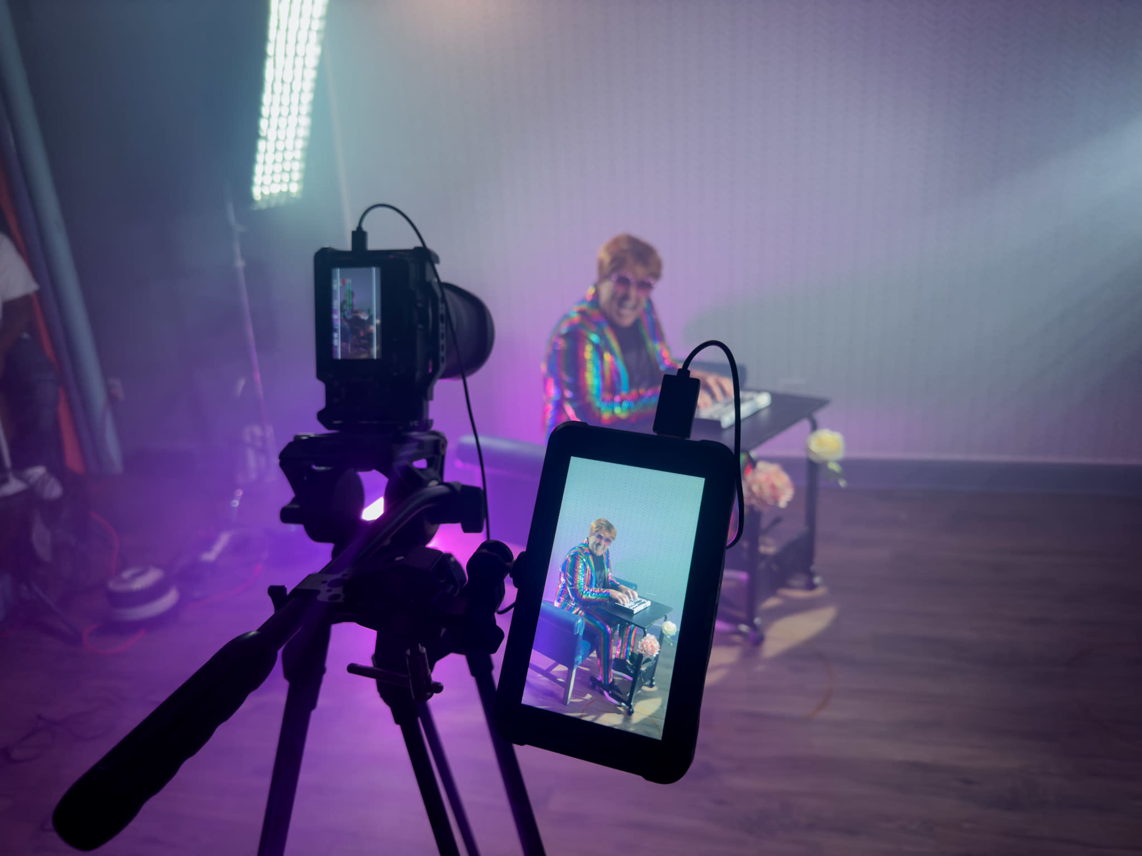 A person is sitting at a desk under colorful lighting, while a camera on a tripod captures the scene on a monitor.