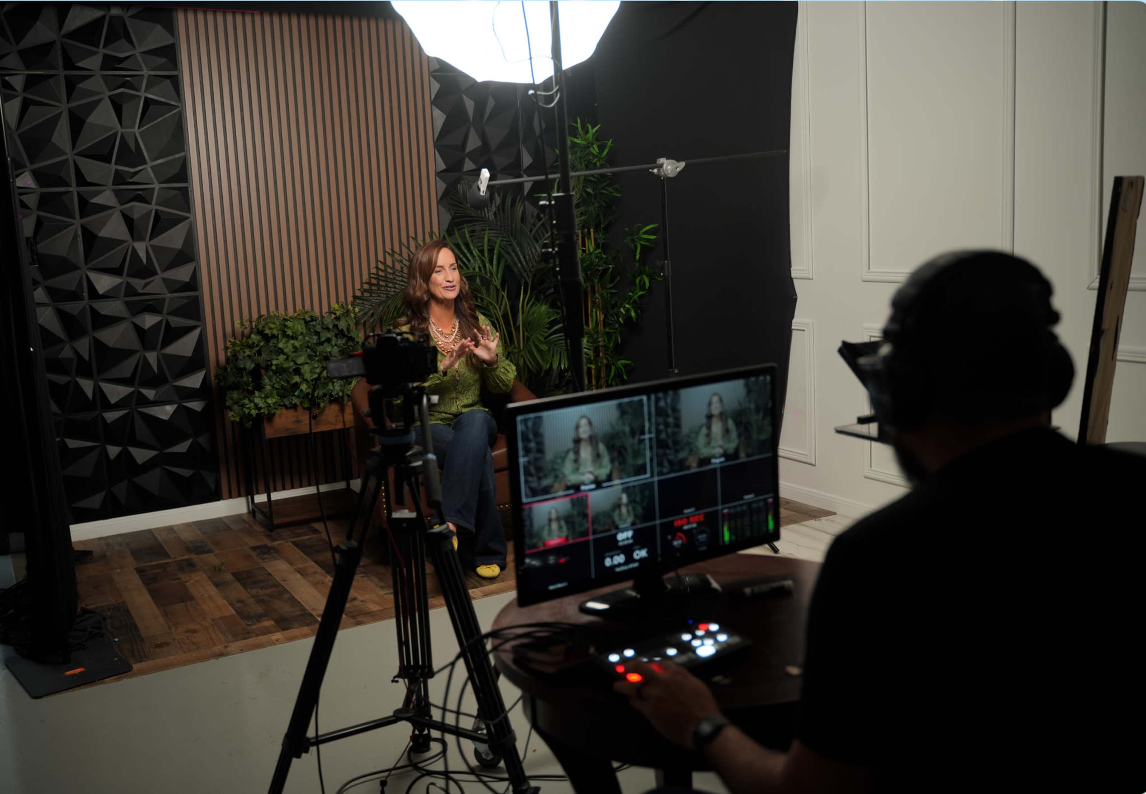A woman is seated in front of a camera in a studio setting, while a person is monitoring the recording from a nearby console.
