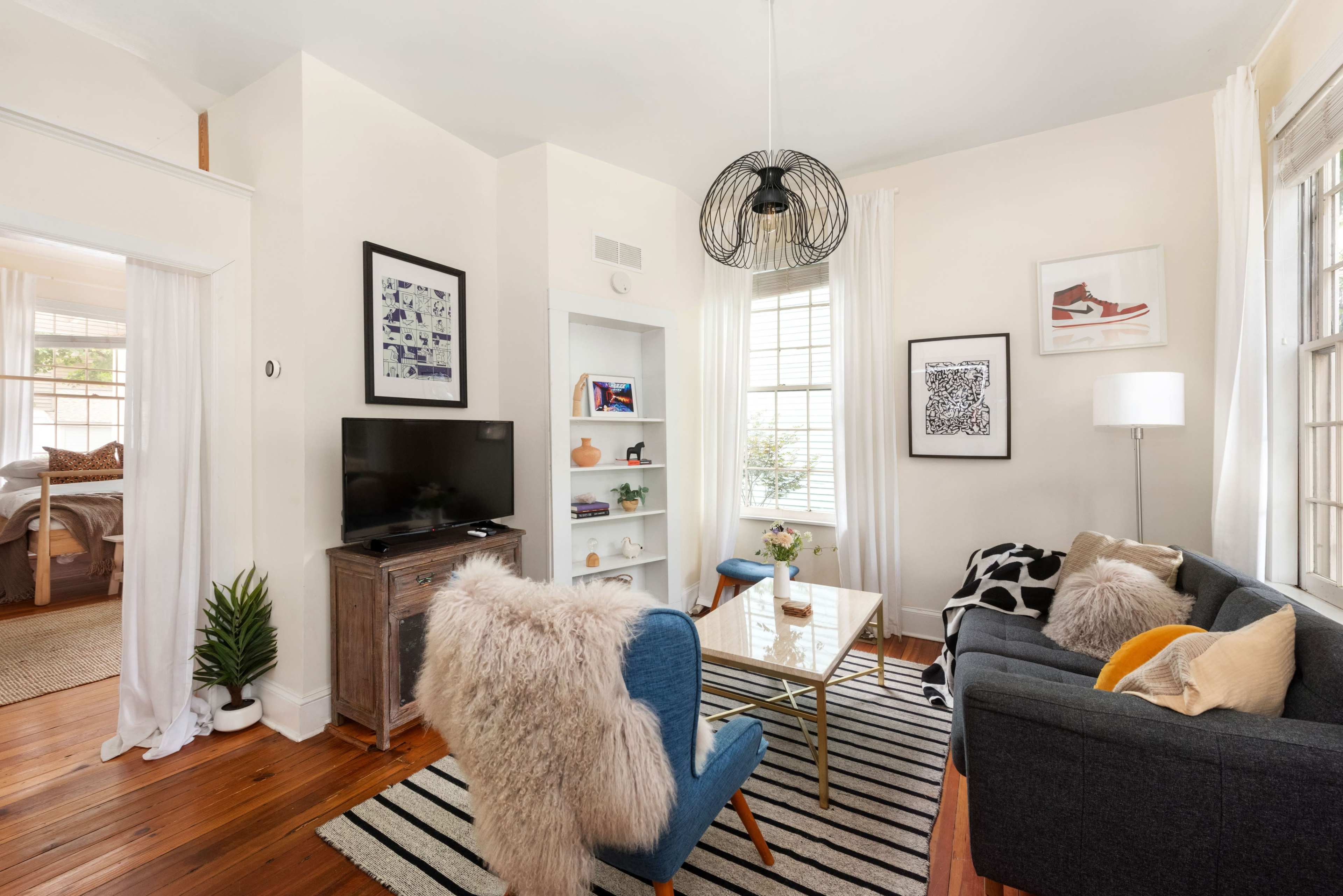 The image shows a stylish living room with a black sofa, a glass coffee table, a TV, and decorative art on the walls, featuring light-colored walls and a striped rug.