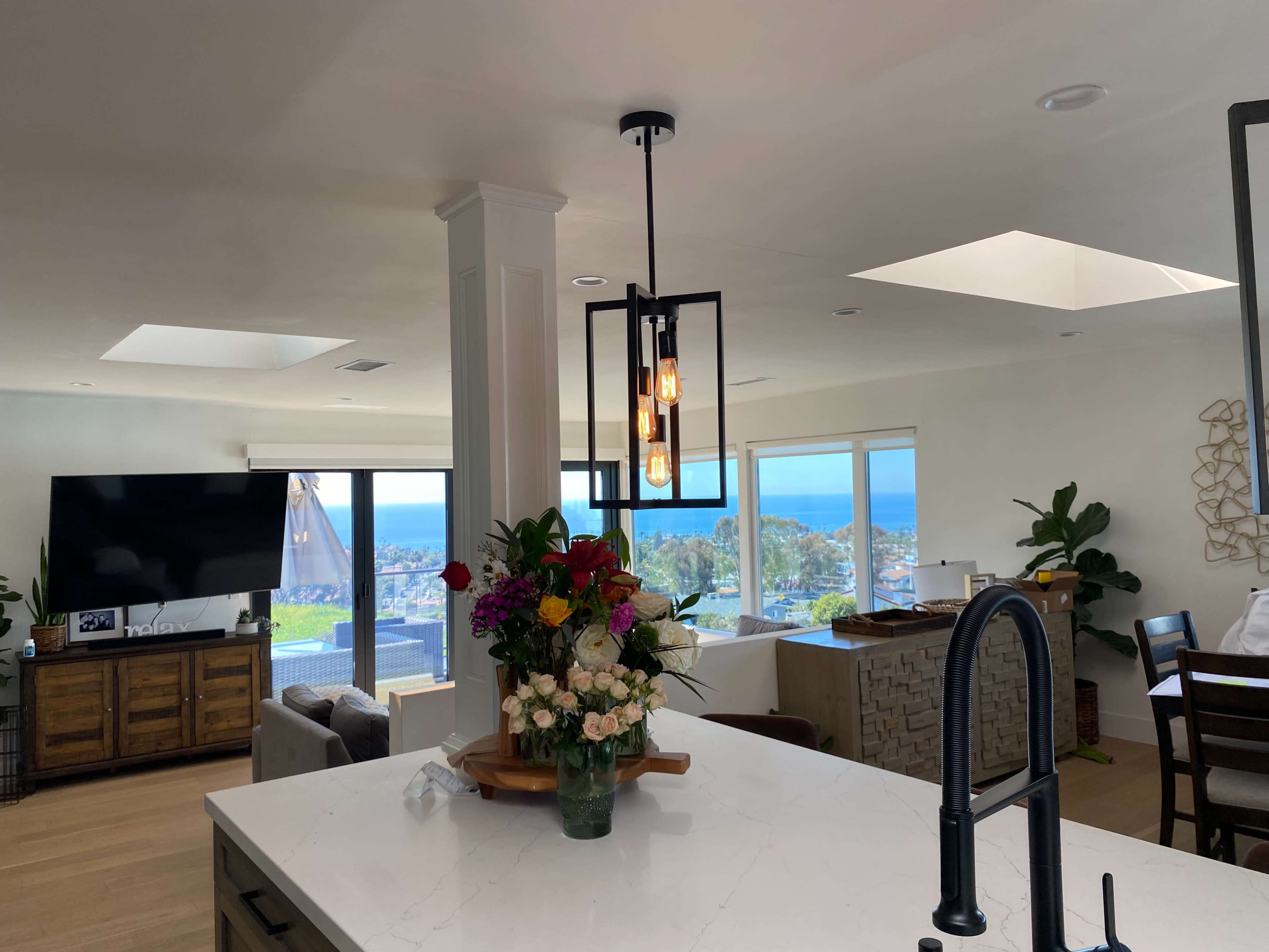 A modern kitchen with a pendant light, a vase of flowers on a countertop, and large windows overlooking a coastal view.