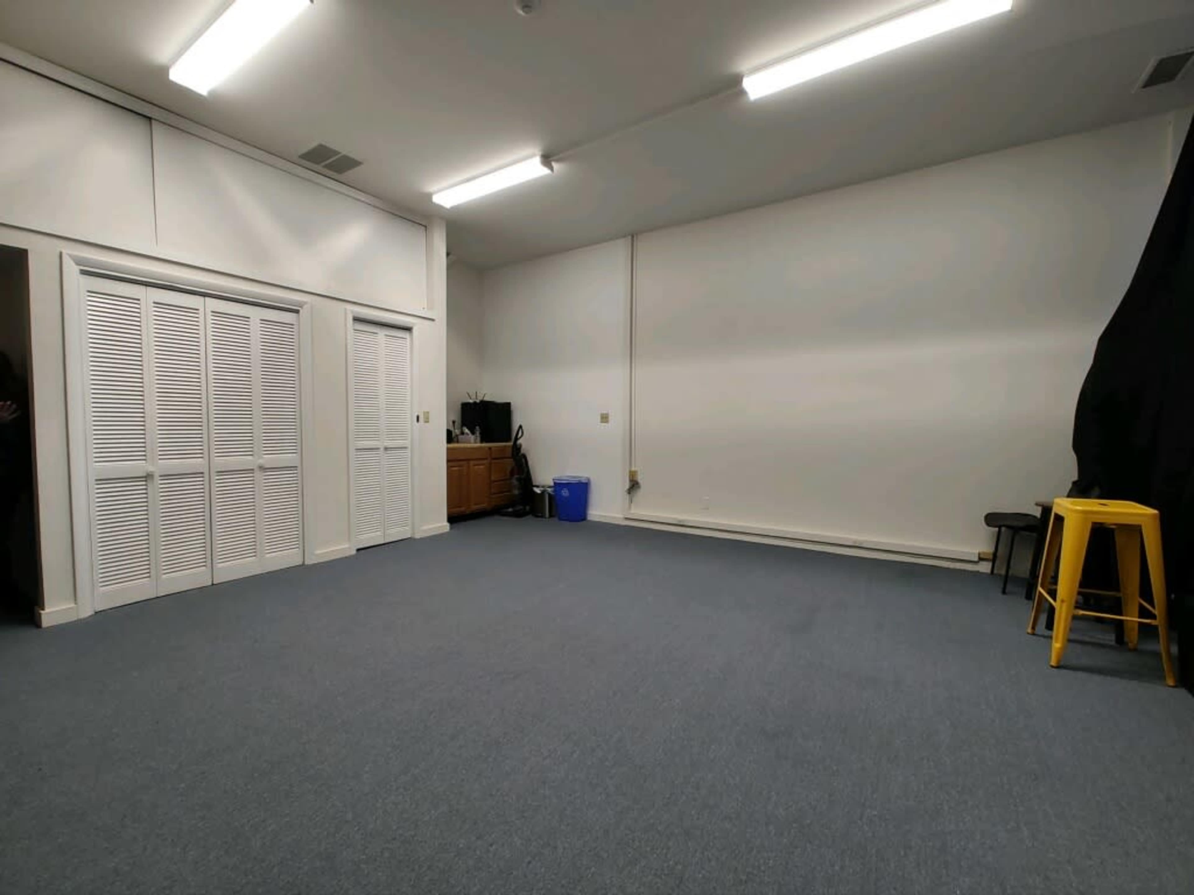 A spacious, empty room with blue carpet, white walls, and minimal furniture including a yellow stool and a blue container.