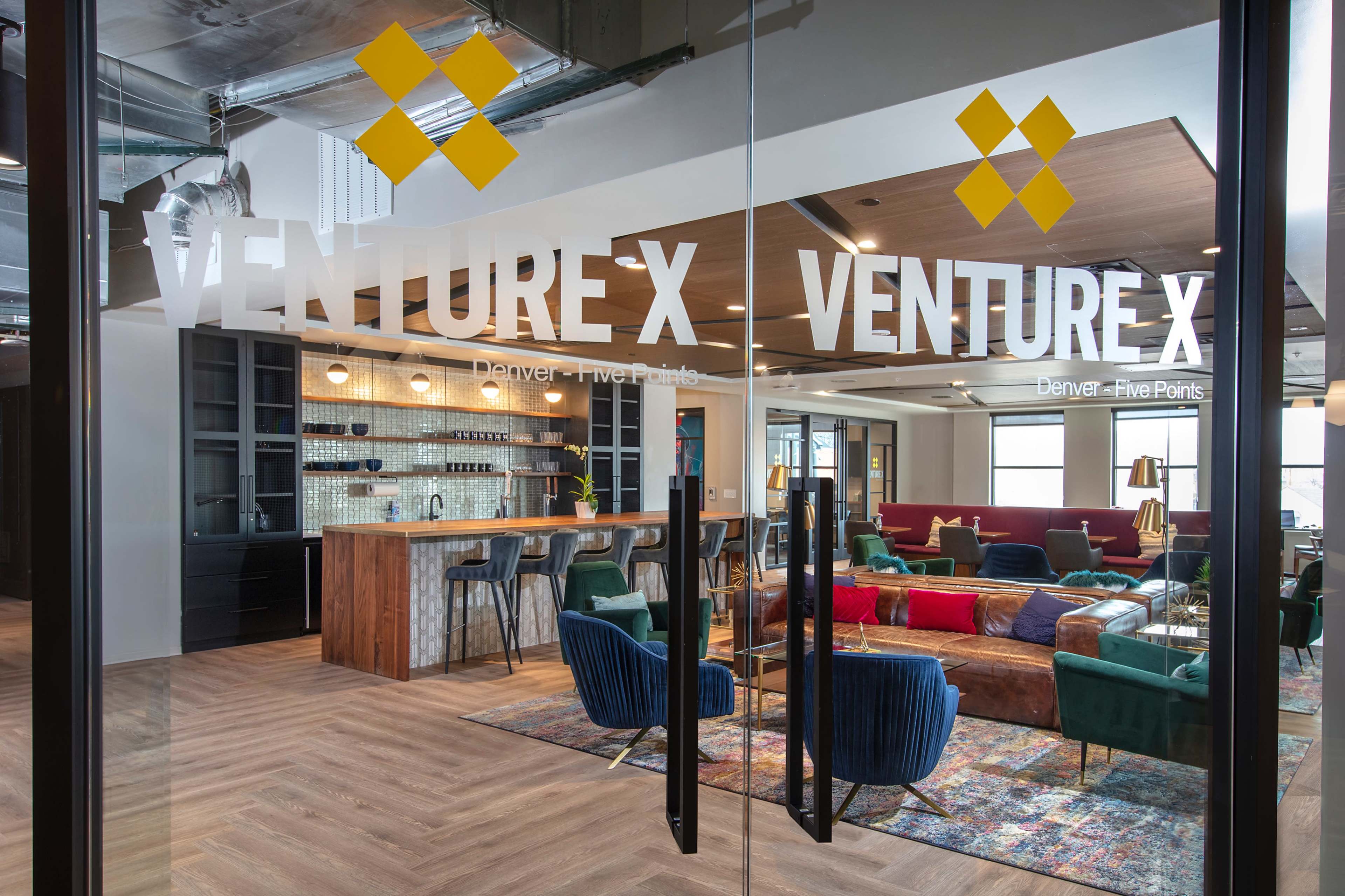 The image shows the entrance to a modern coworking space named Venture X, featuring a spacious interior with a mix of seating areas and a communal kitchen bar.