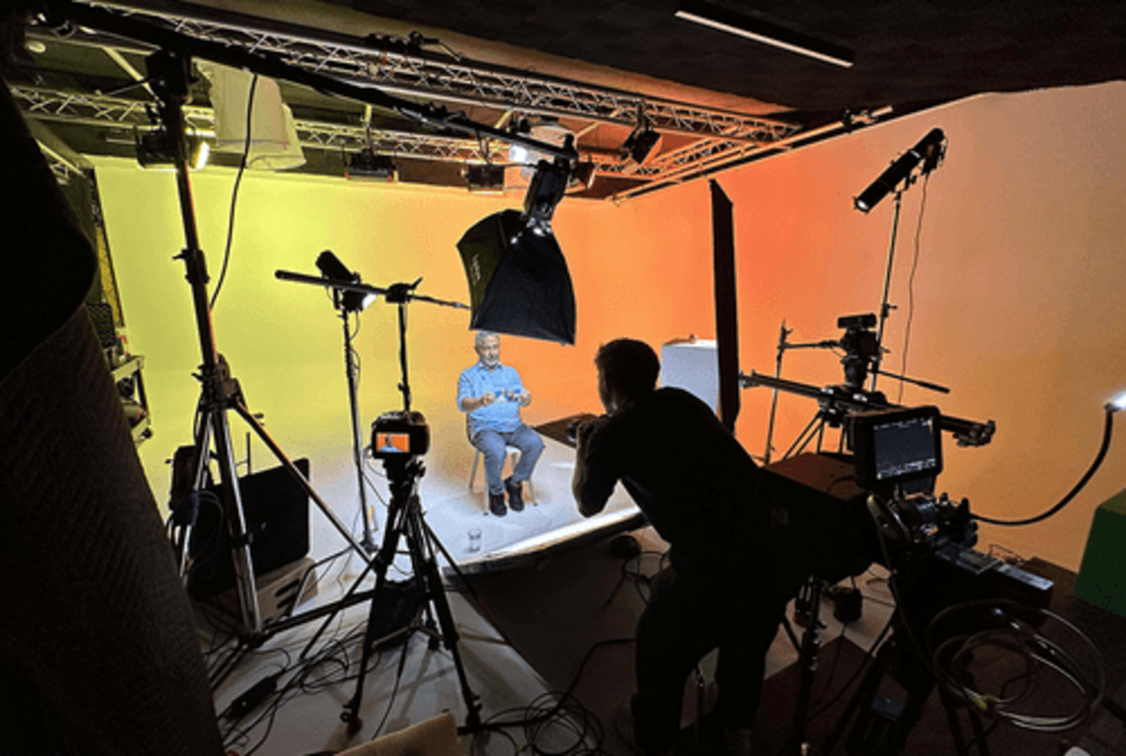 A person is sitting in front of a backdrop in a studio, while another individual captures footage with a camera and various lighting equipment is set up around them.