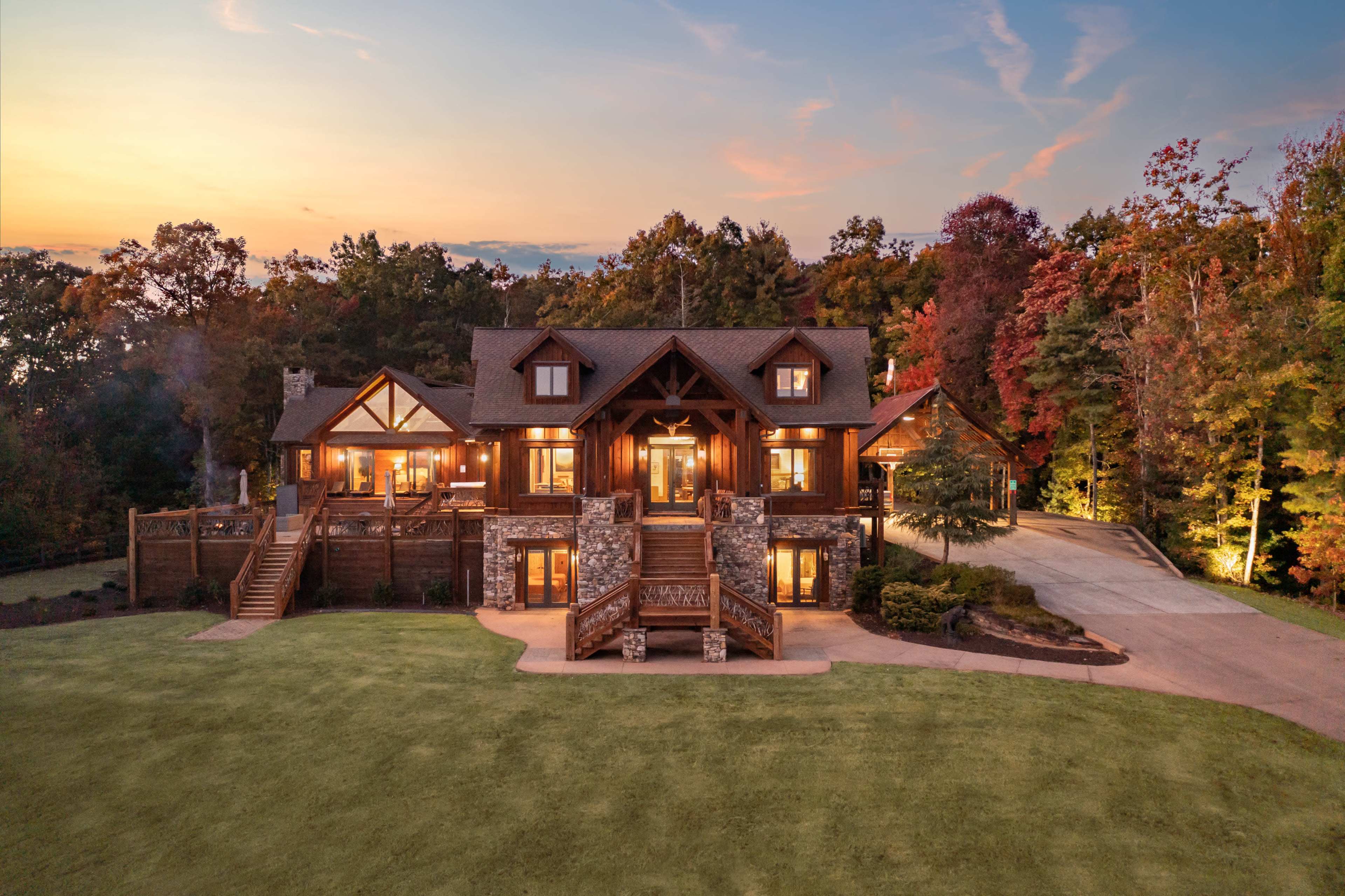 The image shows a large, rustic-style house with a stone and wooden exterior, surrounded by colorful trees and a landscaped yard at sunset.