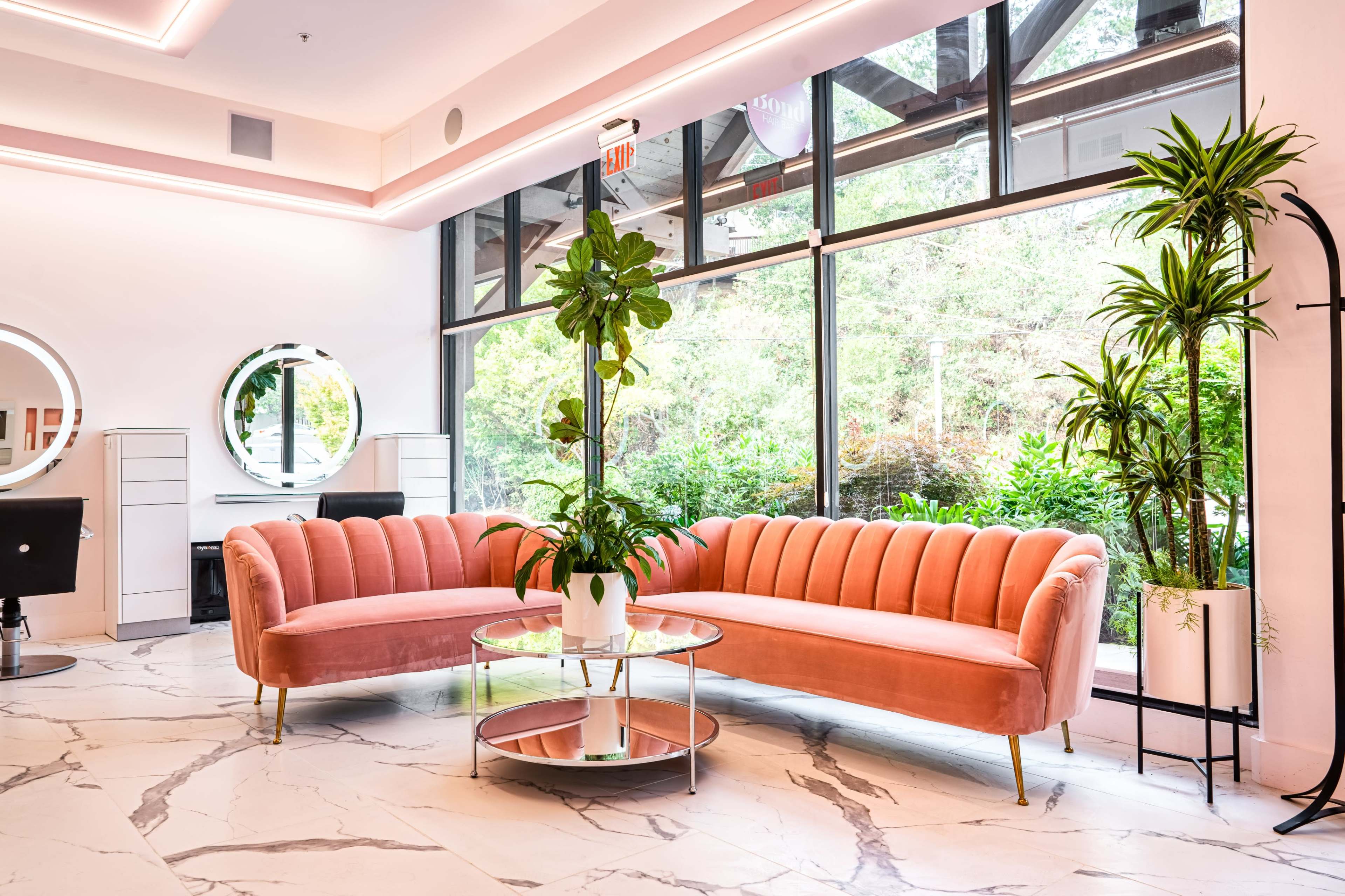 A modern interior with a pink curved sofa, a glass coffee table, and large windows that provide views of greenery outside.