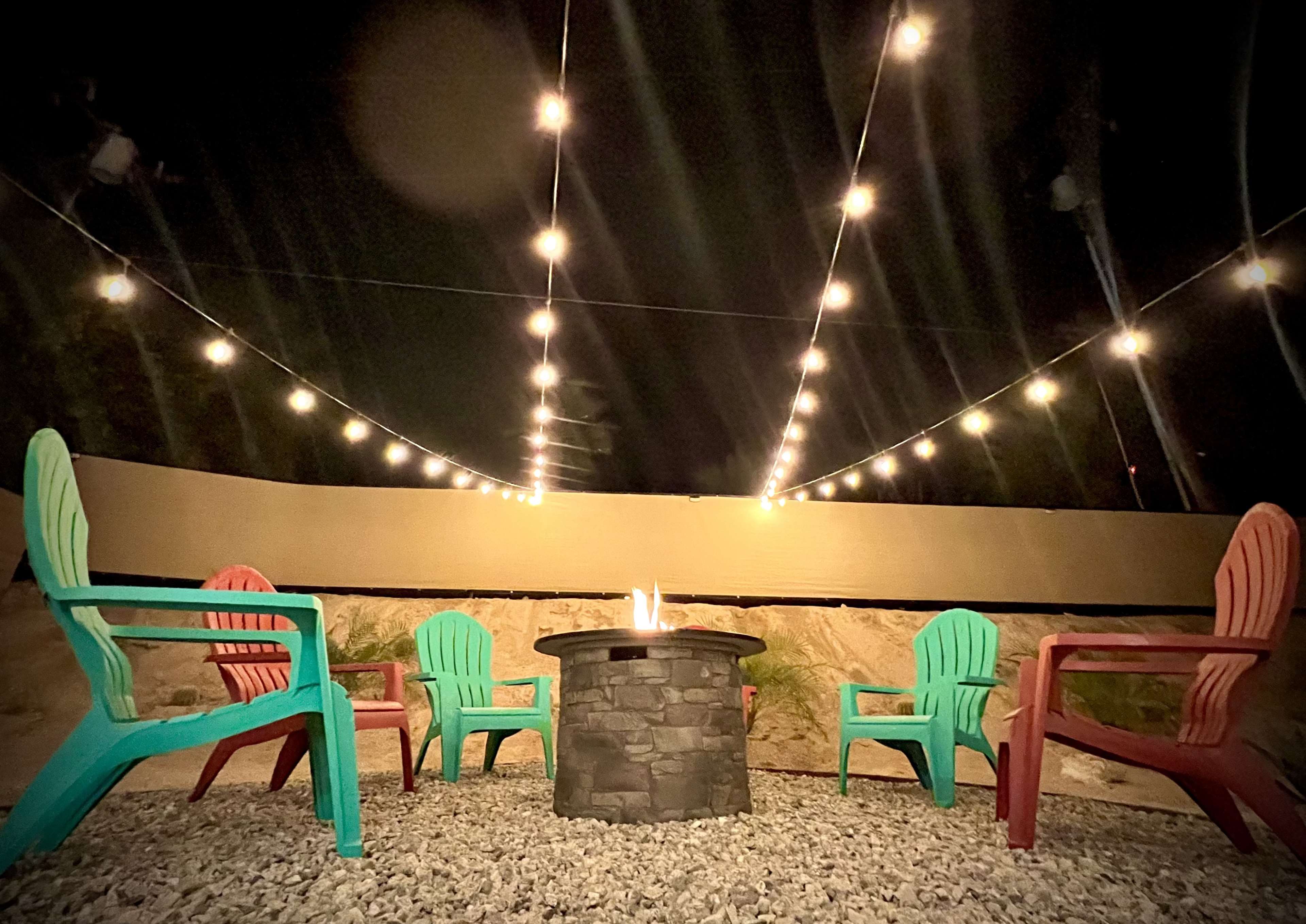 A circular stone fire pit is surrounded by colorful adirondack chairs under a string of lights at night.