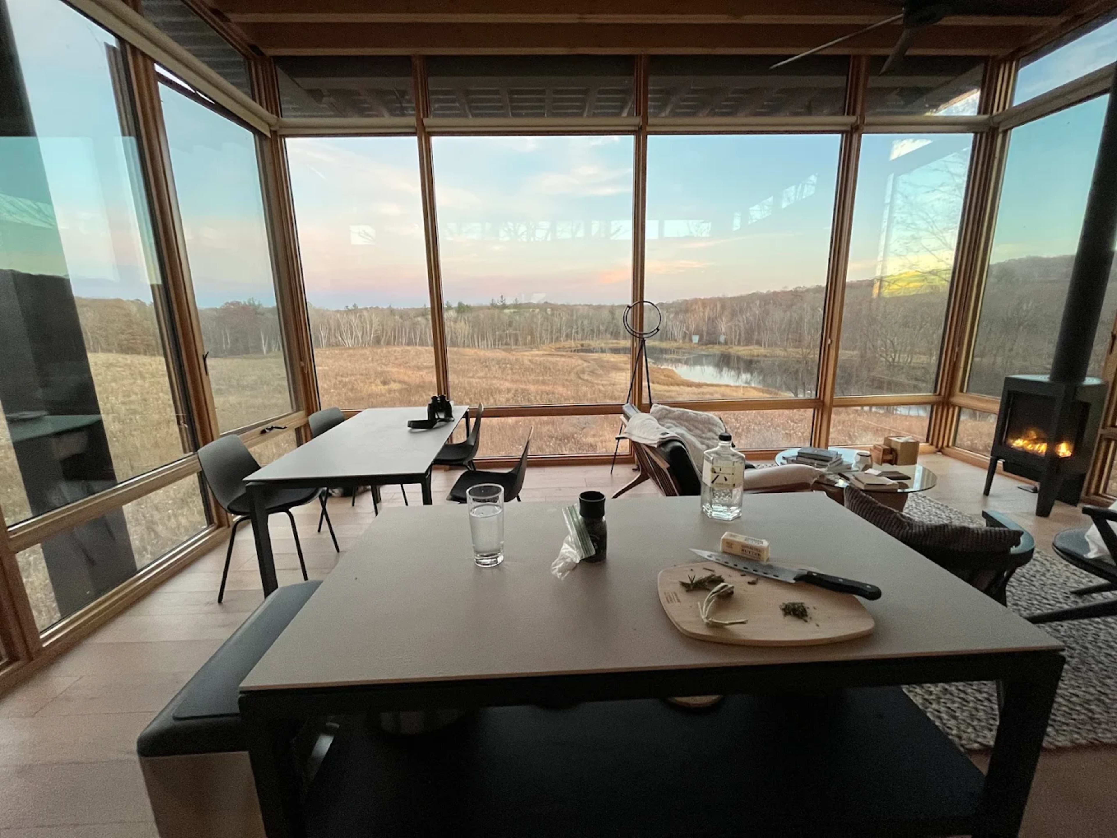 The scene shows a modern dining area with large windows overlooking a grassy landscape and a river.