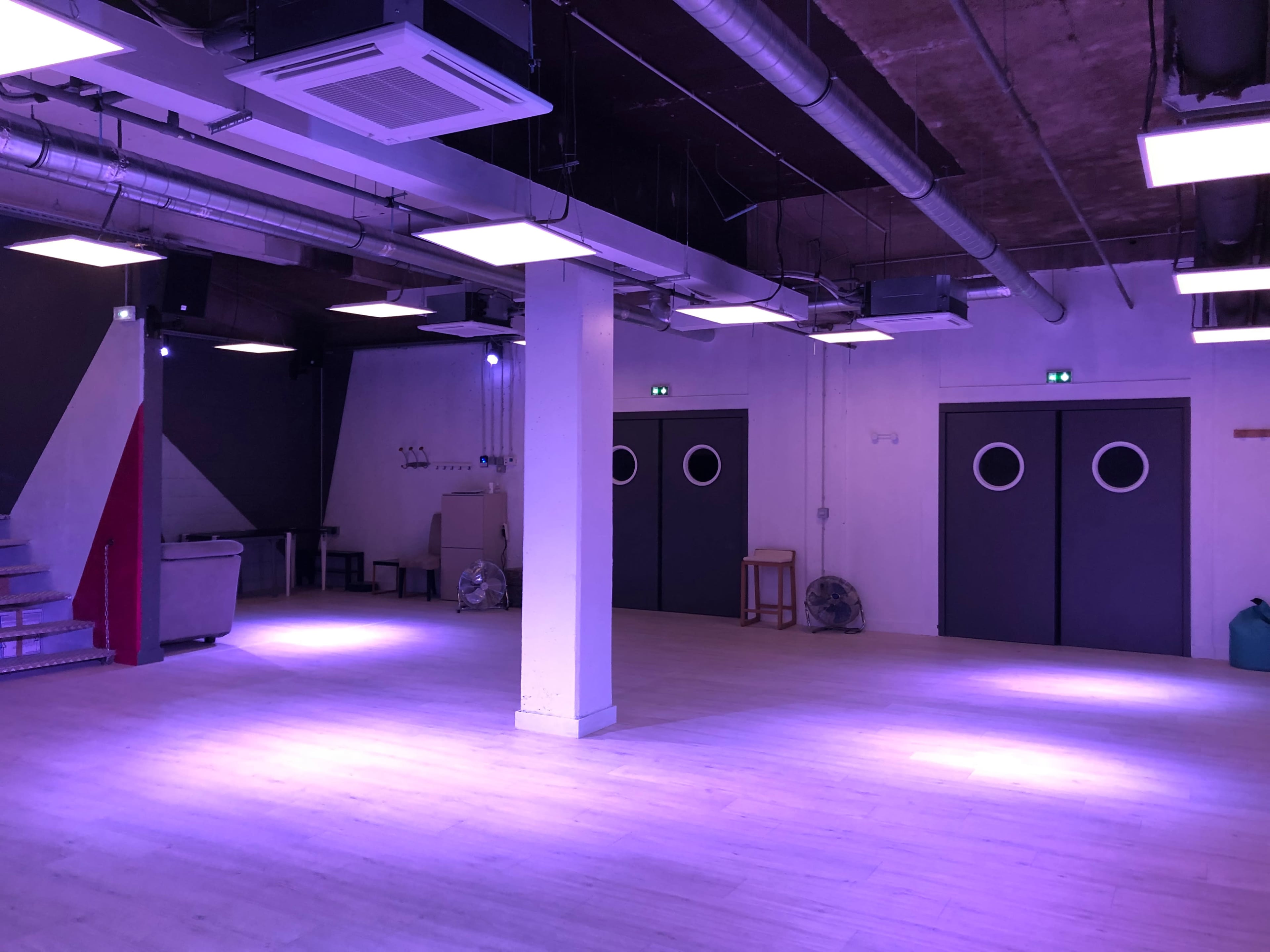 An empty room with wooden flooring, industrial-style ceiling vents, and two closed doors featuring round windows.