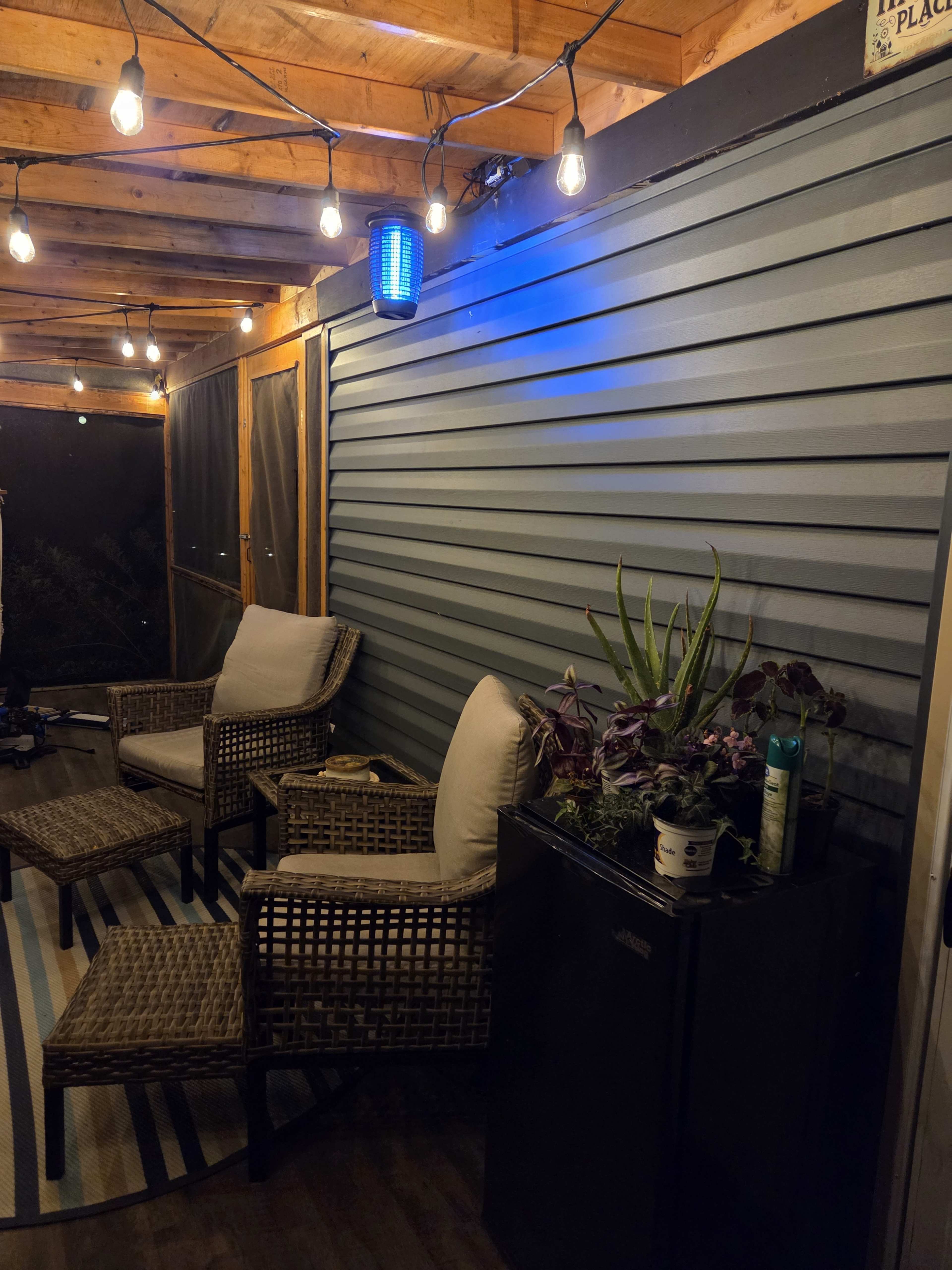 A screened-in porch features two wicker chairs, a small refrigerator, and a wall mounted blue light, illuminated by string lights overhead.