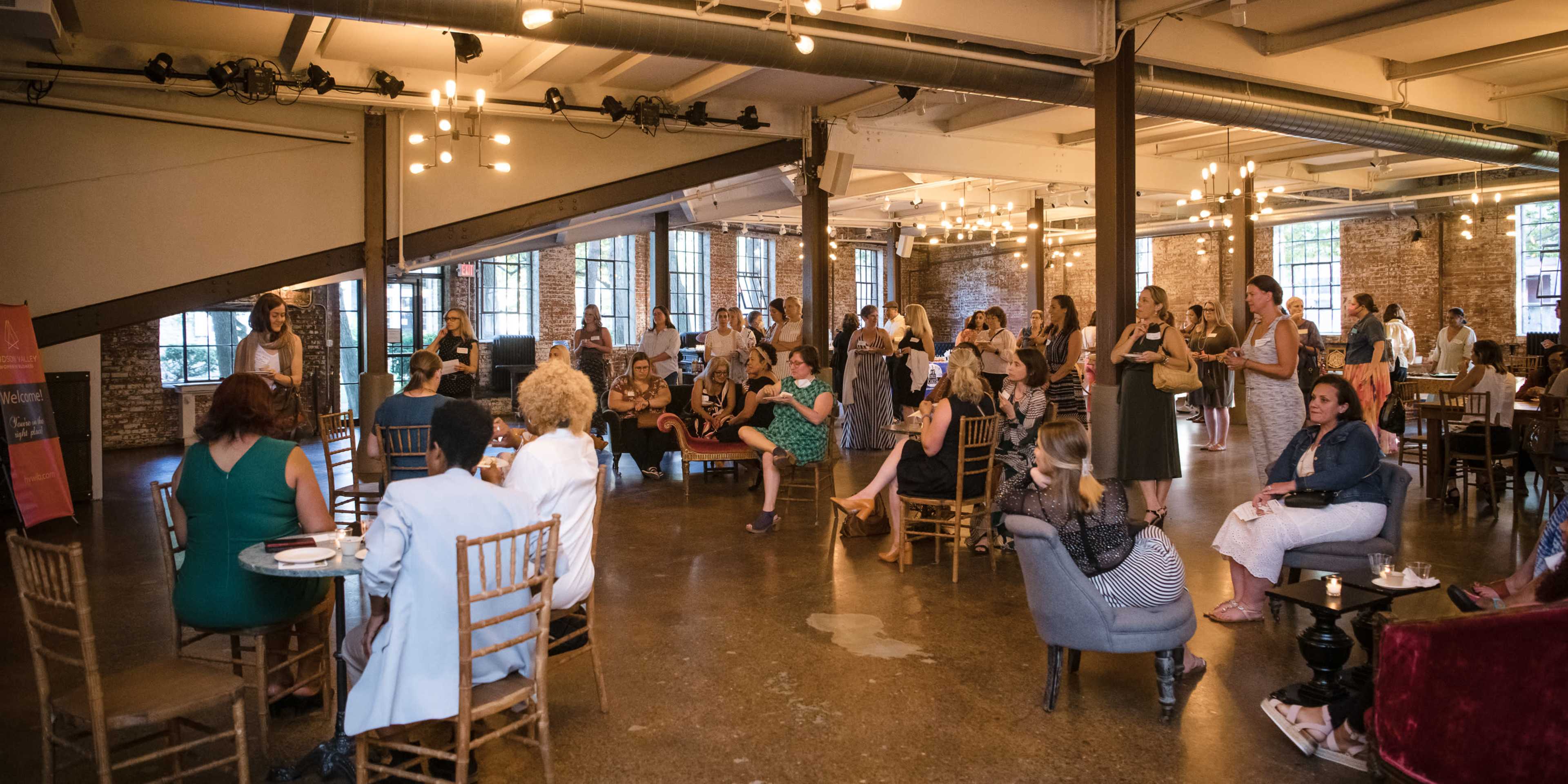A crowd of people gathers in a spacious, well-lit venue with an industrial aesthetic and large windows.