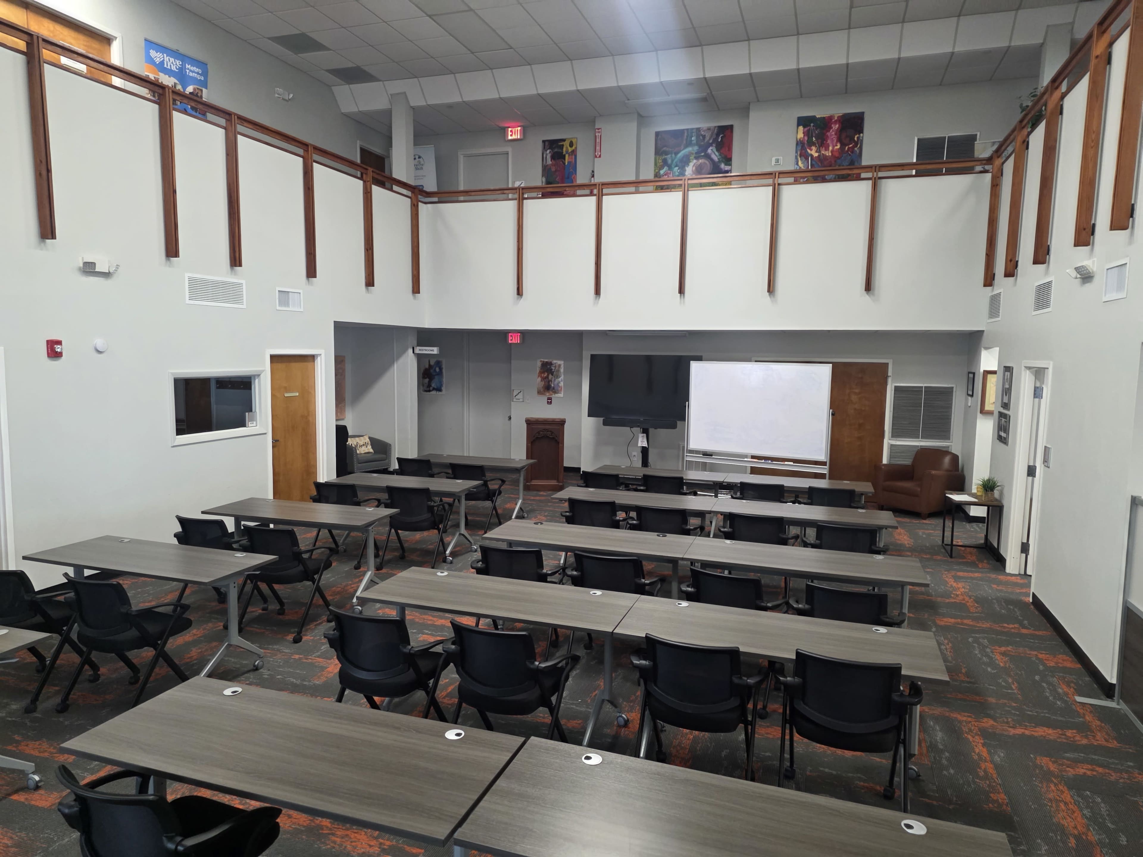 The image shows a spacious meeting room with multiple rows of tables and chairs, a large screen, and a presentation area, illuminated by overhead lights.