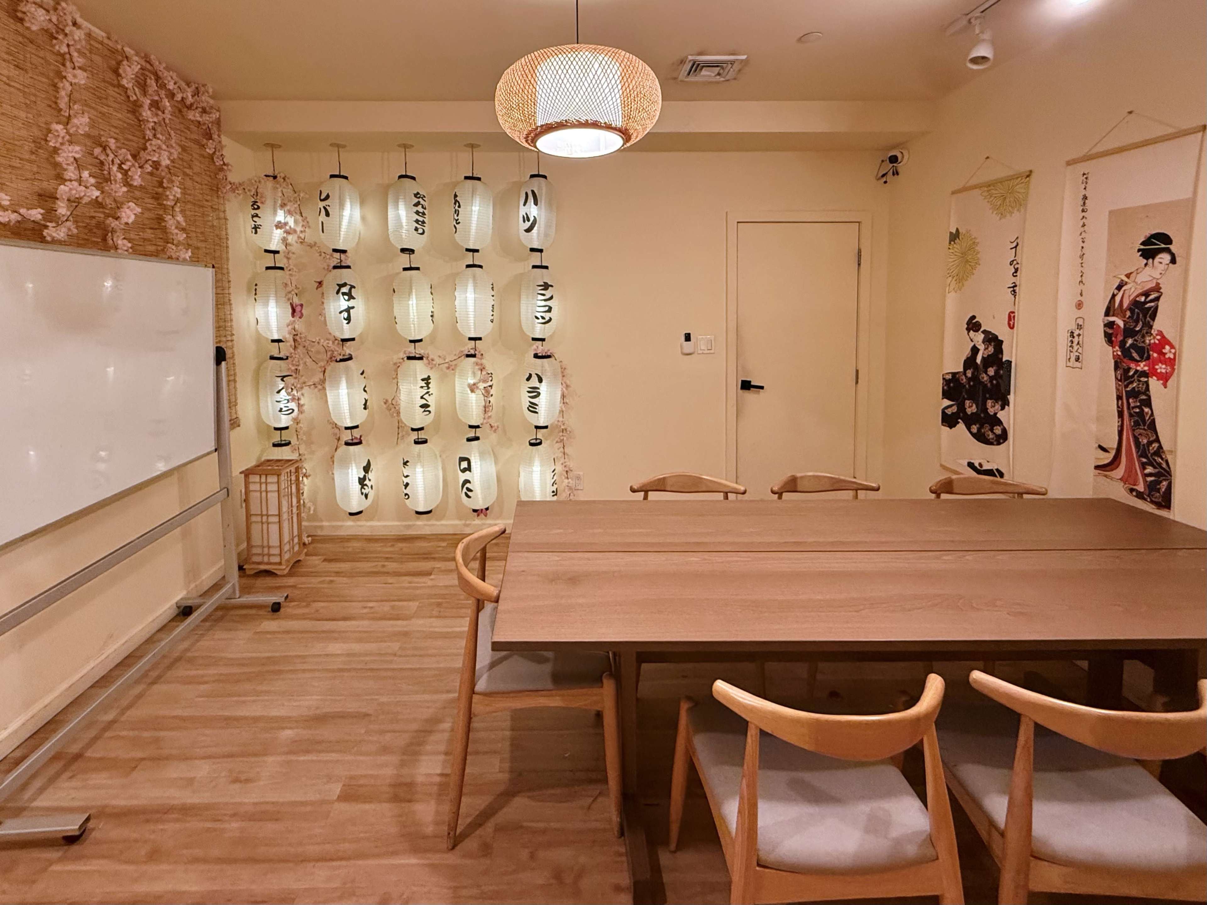 The image shows a cozy room with wooden flooring, a long dining table surrounded by wooden chairs, and a wall adorned with illuminated lanterns and traditional artwork.