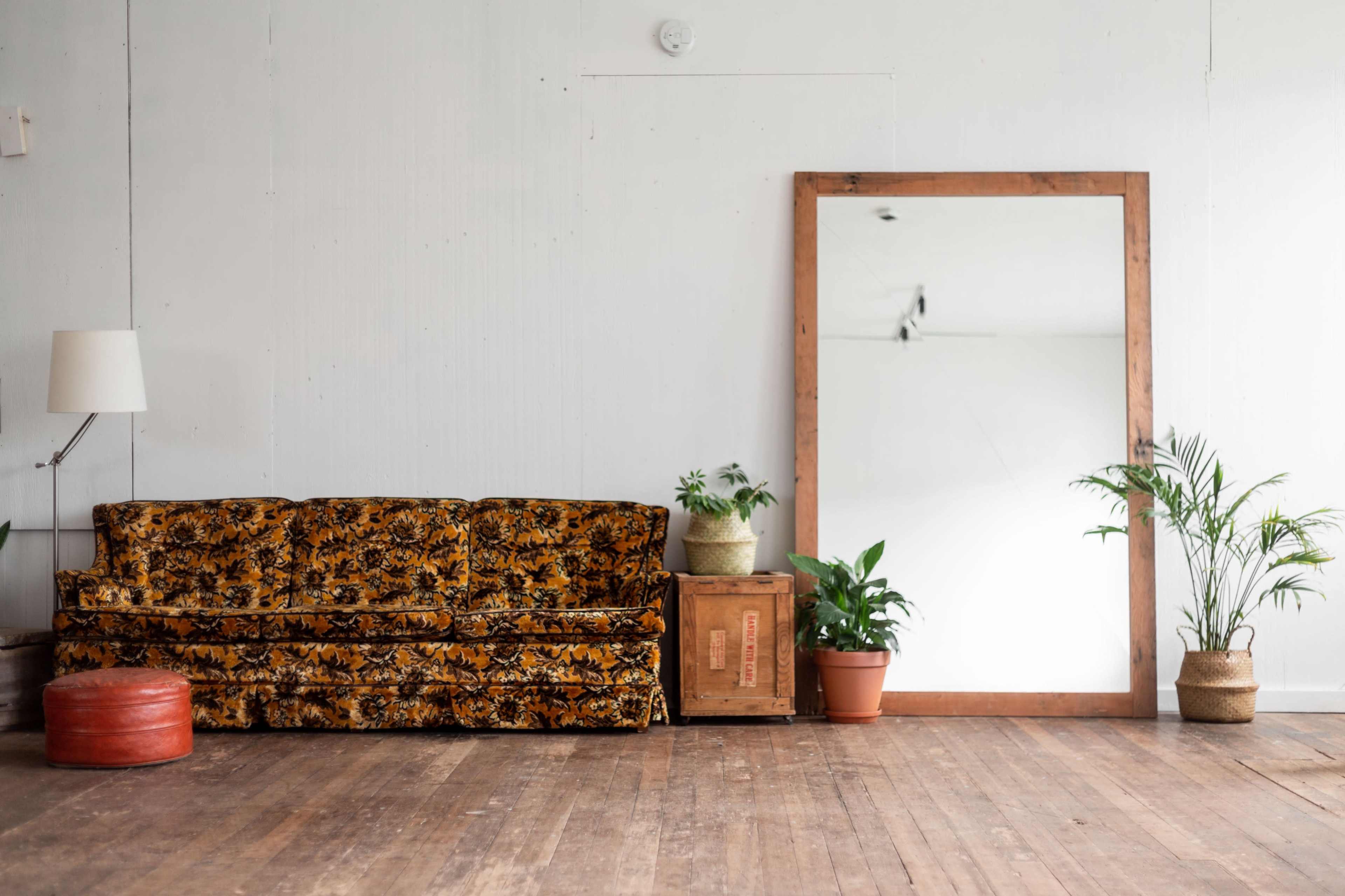 A vintage floral couch sits next to a large mirror and potted plants in a minimally decorated room with wooden flooring.