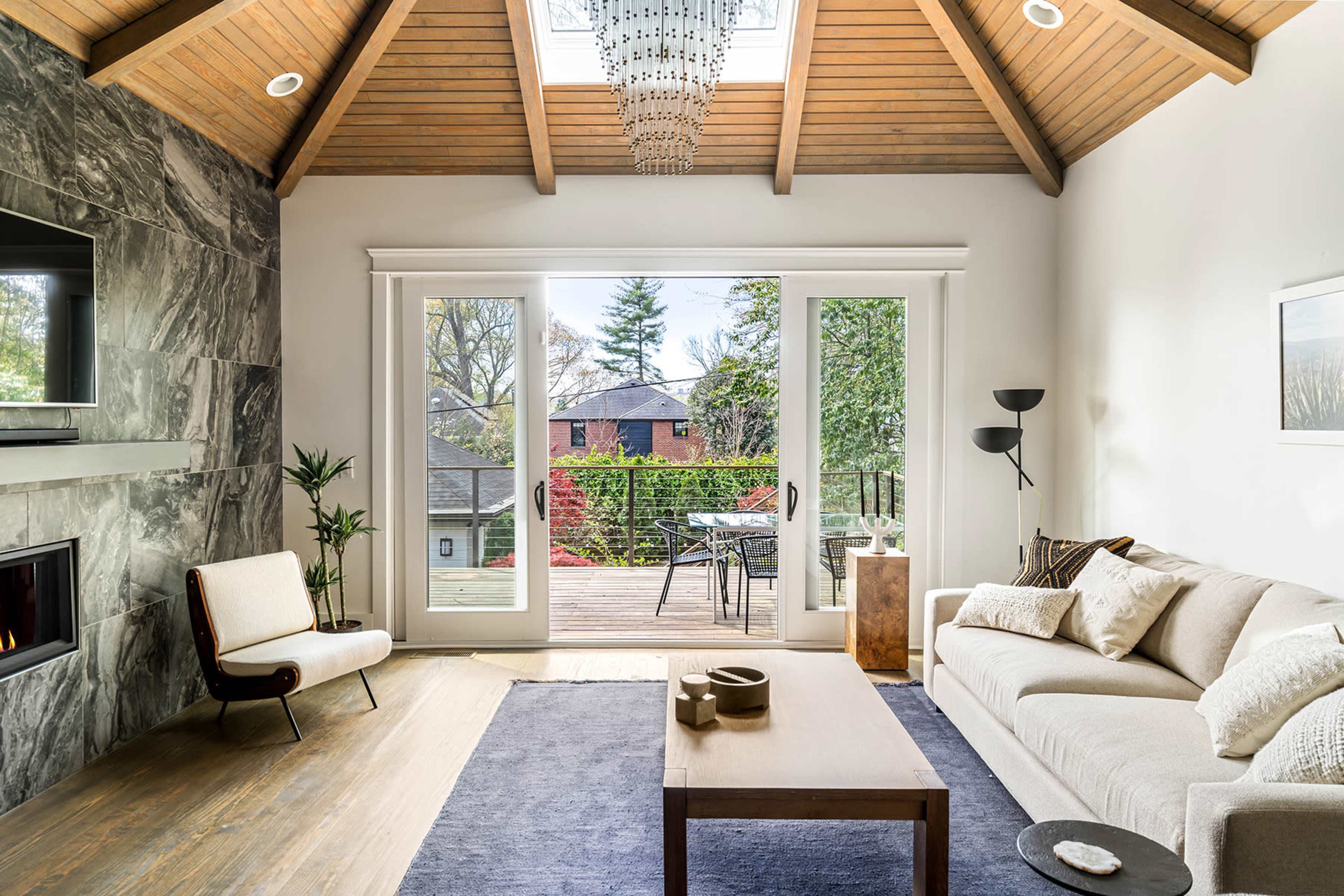 A modern living room with a high wooden ceiling, a large window opening to an outdoor deck, and minimalistic furniture.