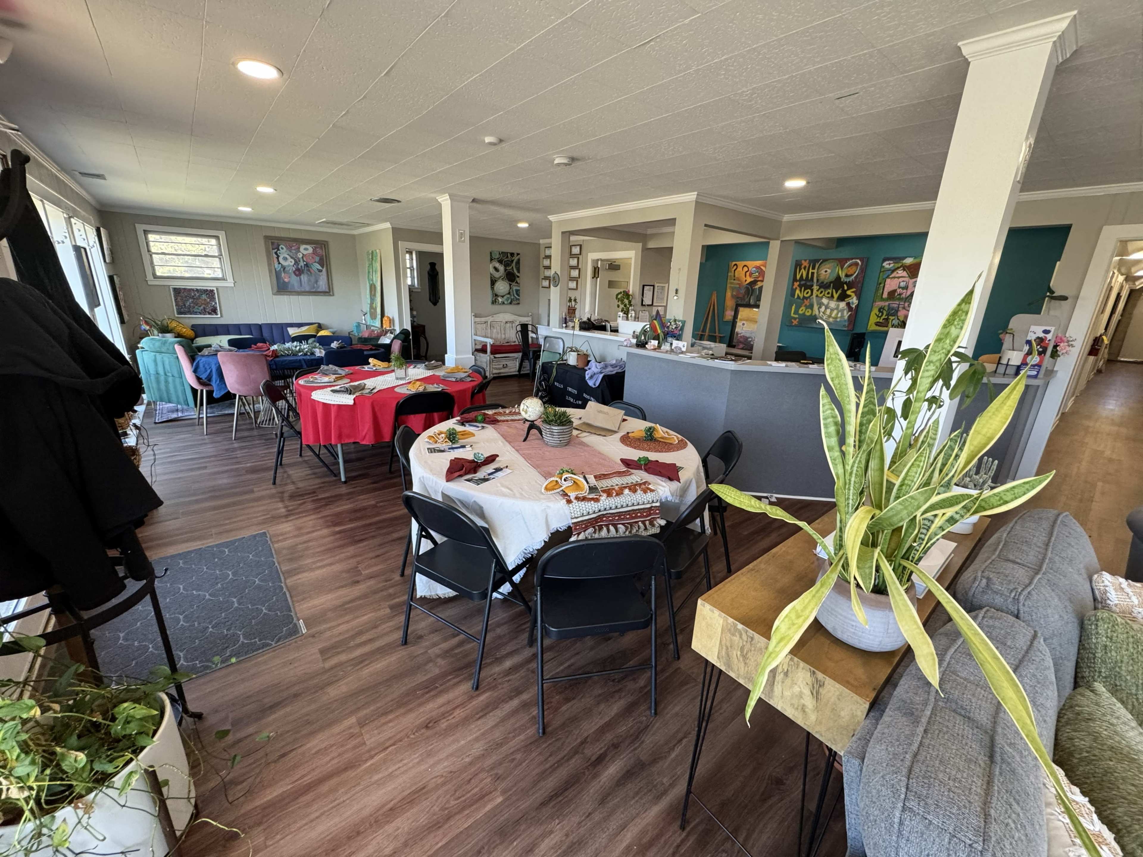 The image shows a spacious, brightly lit community room furnished with tables covered in tablecloths and surrounded by chairs, with colorful artwork decorating the walls.
