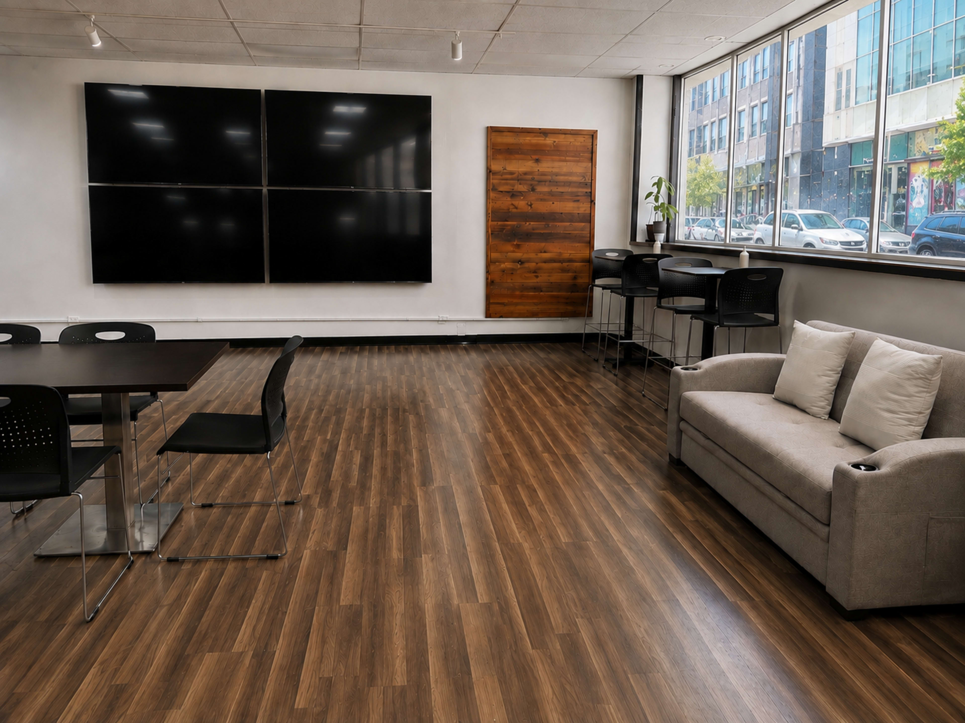 The image shows a modern, open space featuring a seating area with a sofa, a table with chairs, and large windows displaying an urban view.
