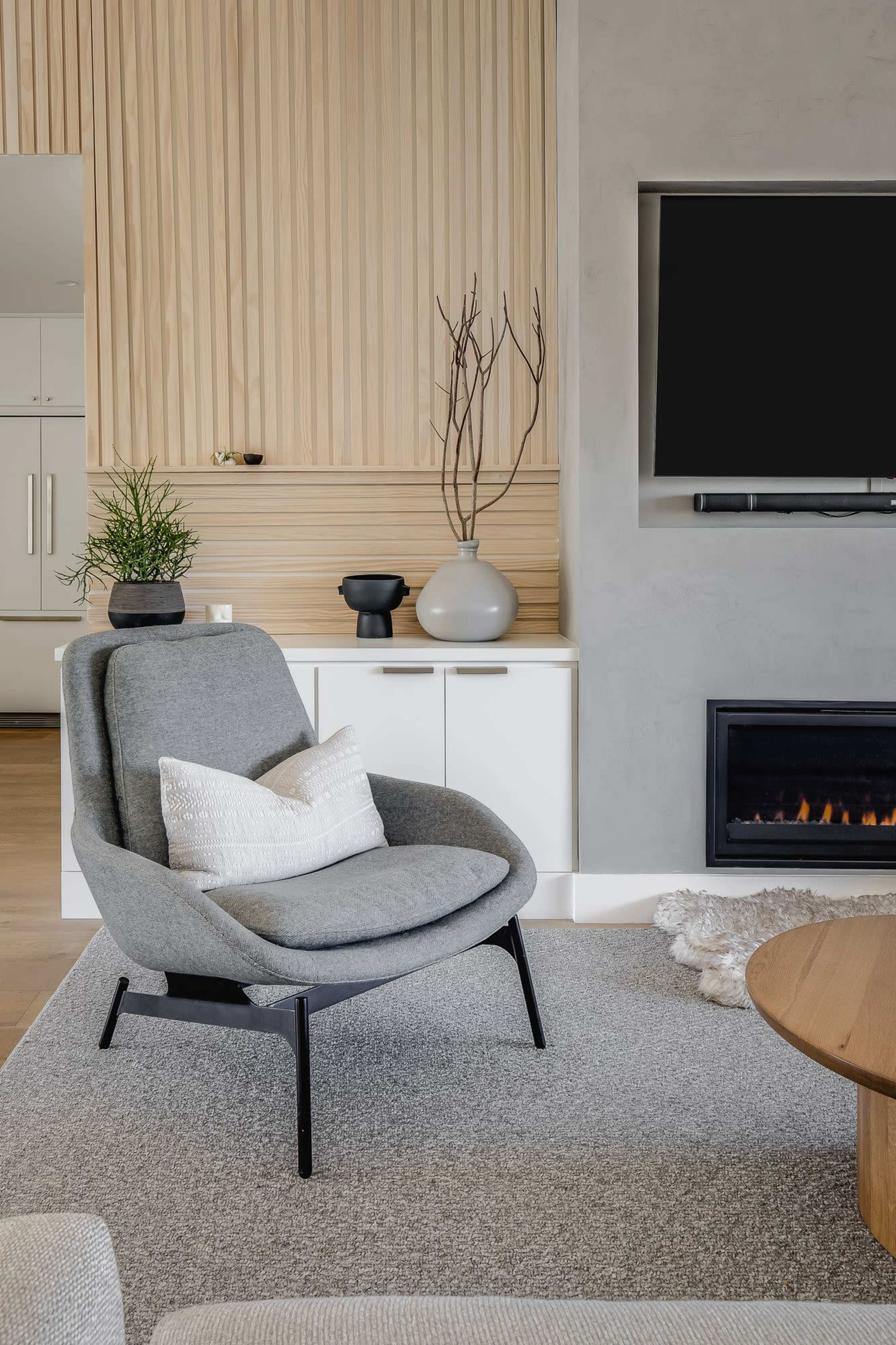 A gray chair with a decorative pillow is positioned next to a wooden table in a modern living room featuring a minimalist design with a TV and a fireplace.