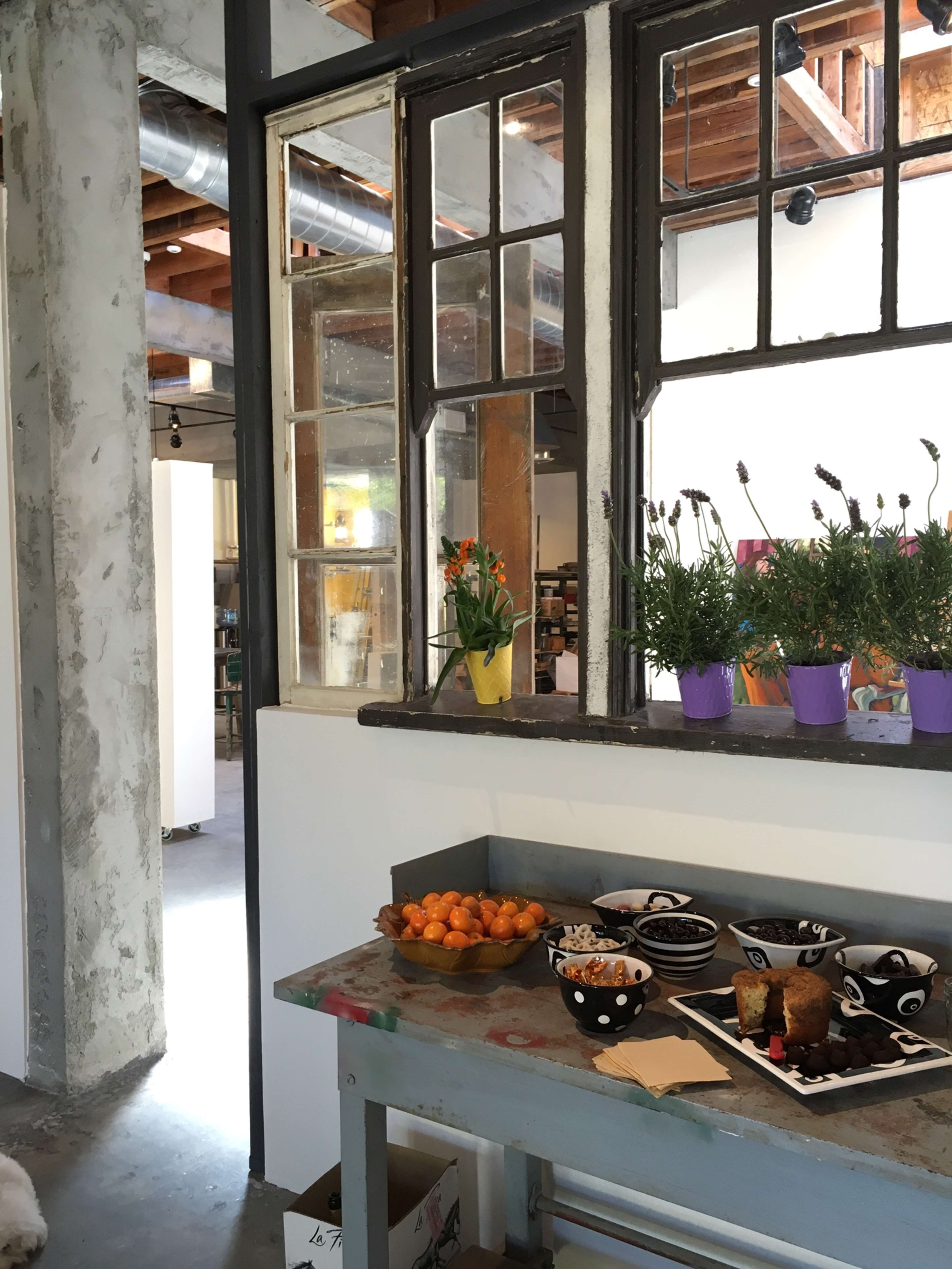 A table with fruit, baked goods, and snacks is set against a backdrop of large windows and potted plants in a modern space.