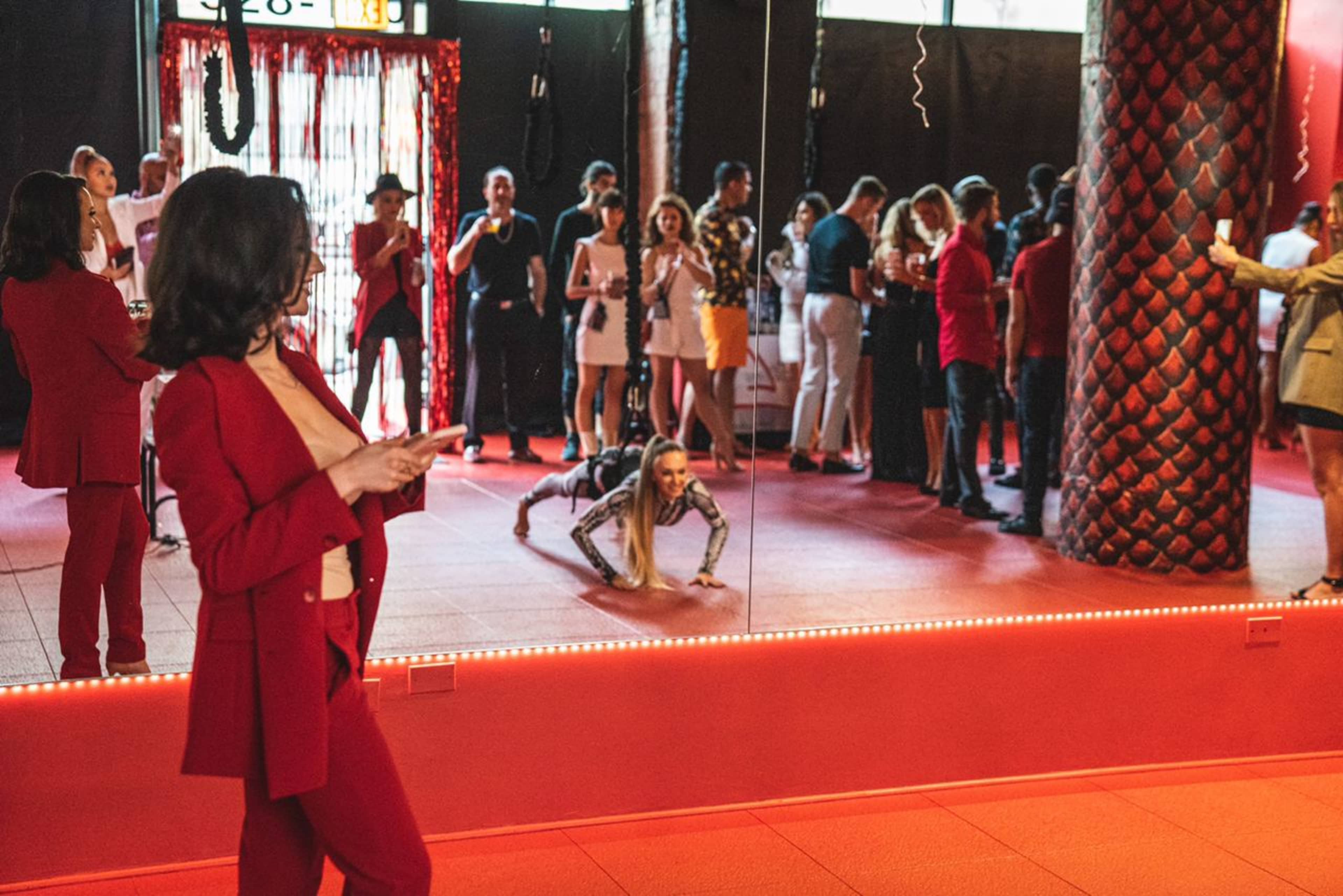 A group of people socialize in a mirrored room, while a woman in a red suit checks her phone and another performs push-ups.