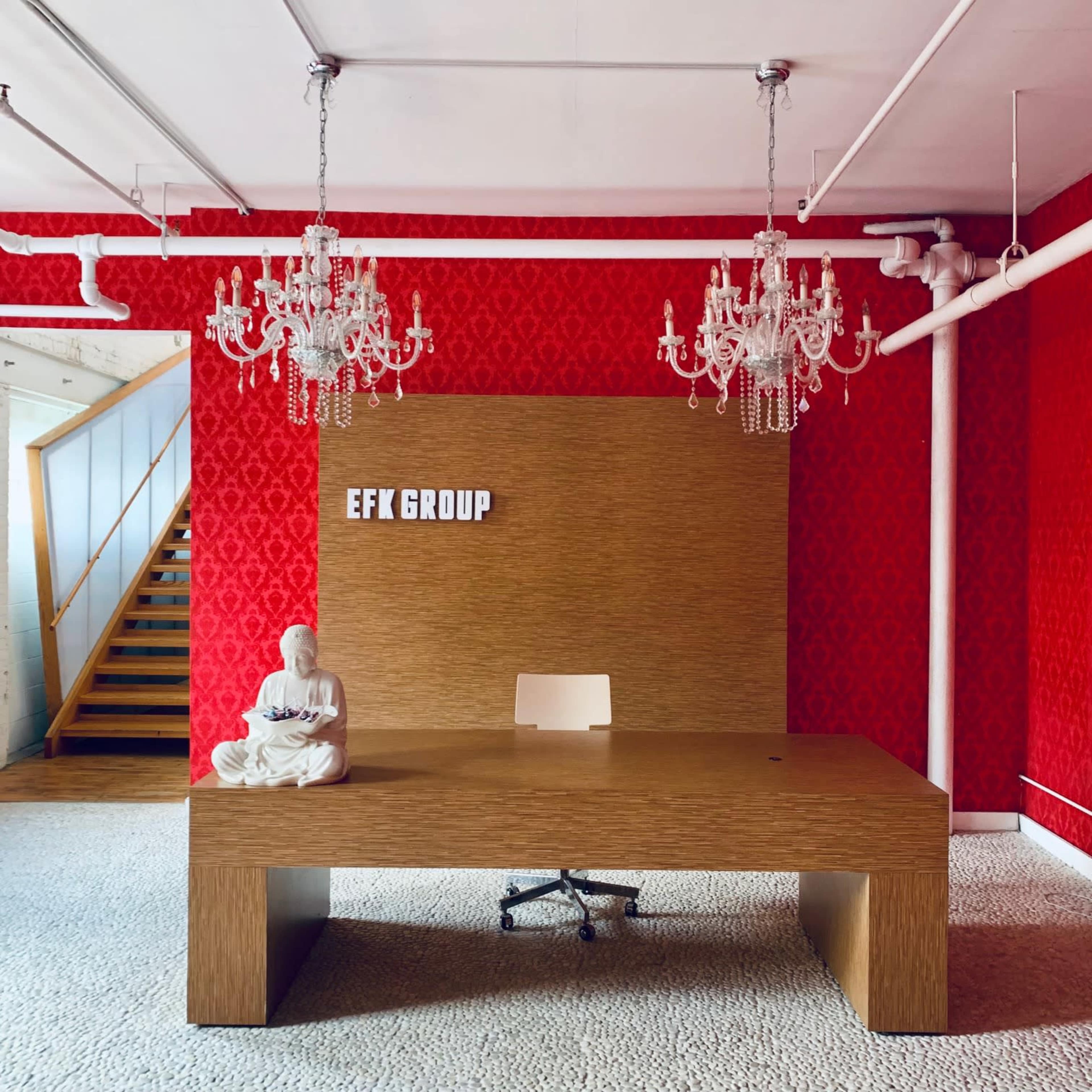 A modern office reception area with a large wooden desk, a white chair, and two ornate chandeliers hanging from the ceiling, set against a striking red patterned wall.