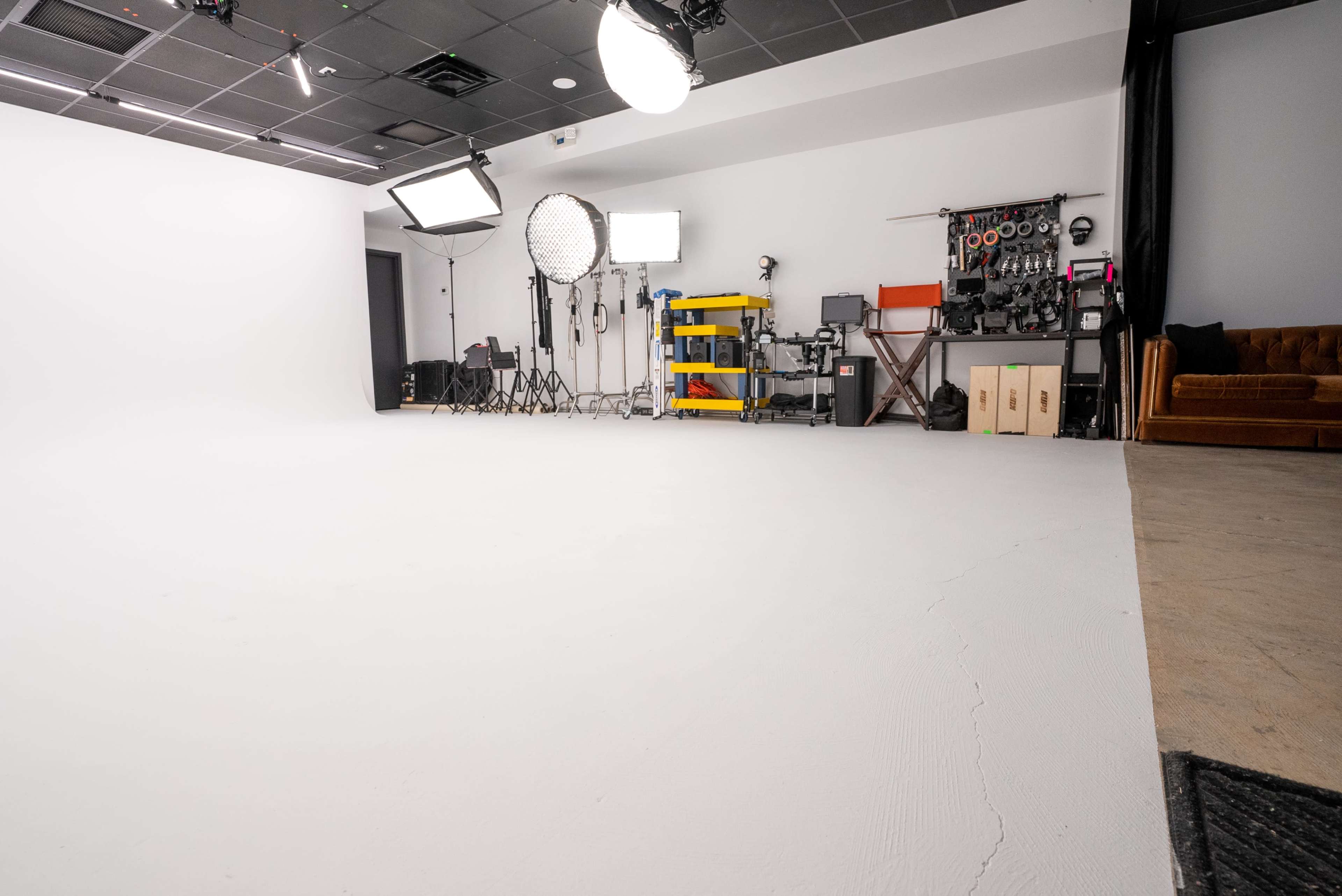The image shows a photography studio with a white backdrop, various lighting equipment, and a brown couch against a wall.