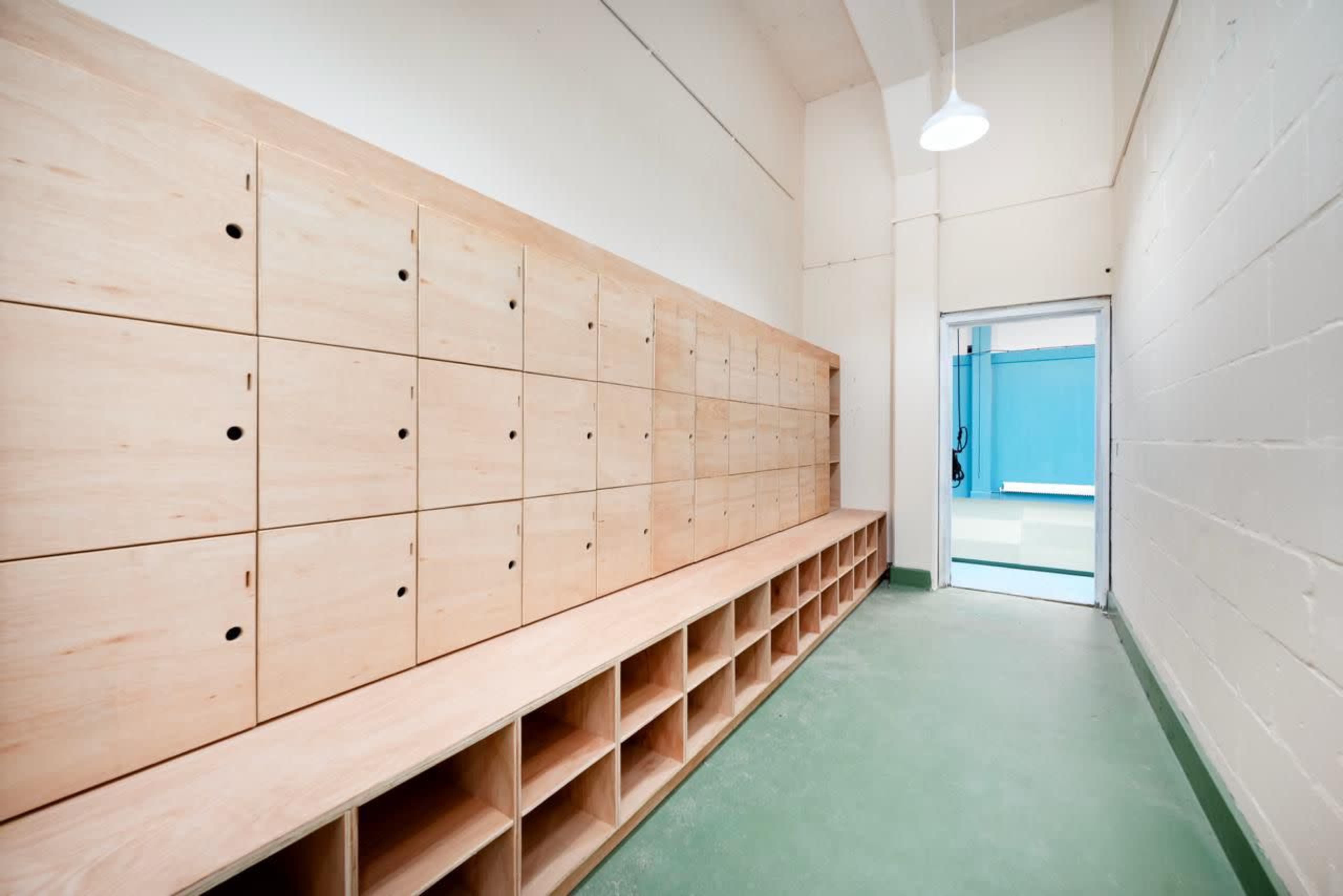 The image shows a narrow hallway featuring a row of wooden lockers on one wall and a doorway leading to a blue-walled room.