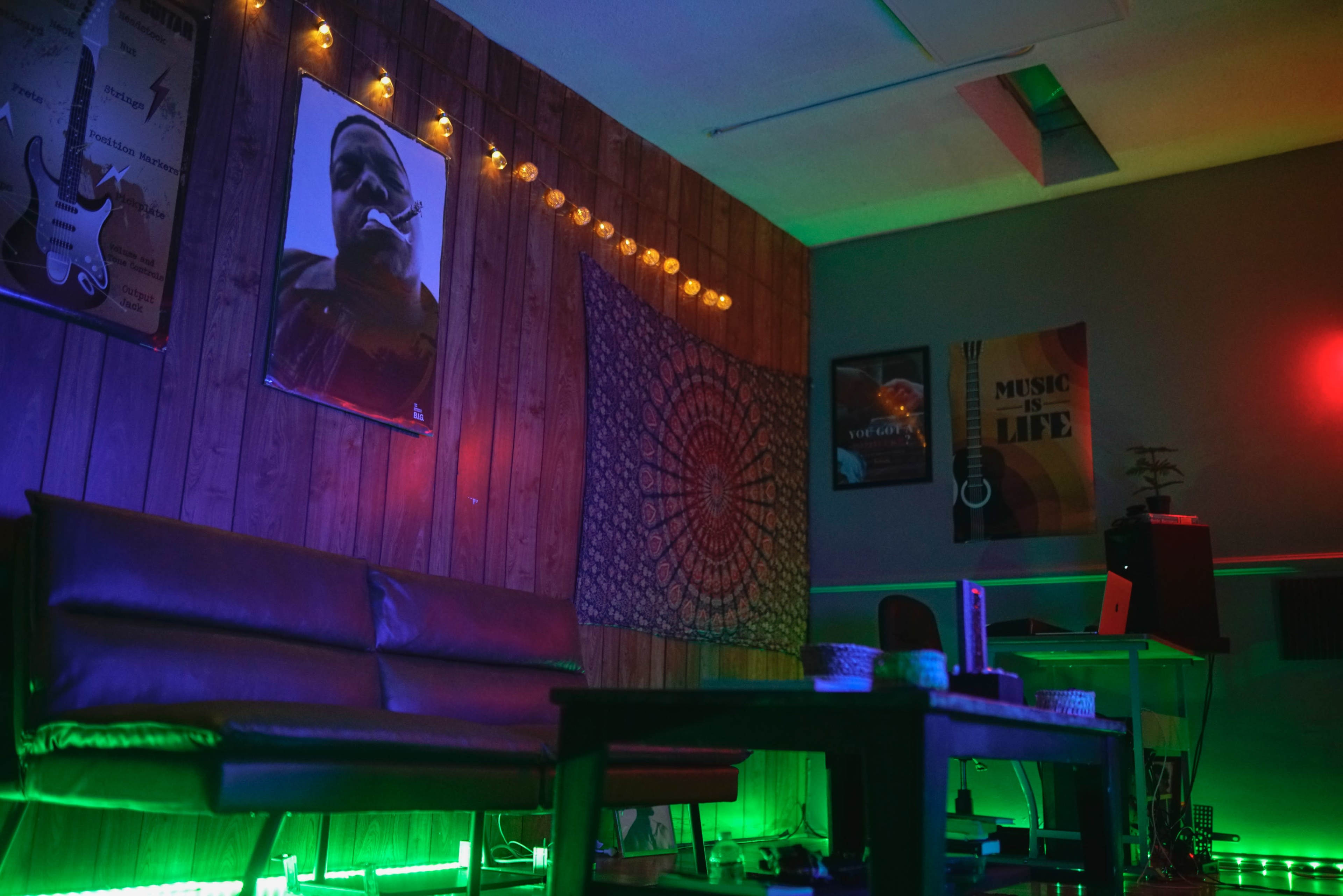 A cozy room features a dark leather sofa, colorful lighting, framed posters on the wooden walls, and a small table in front of it.