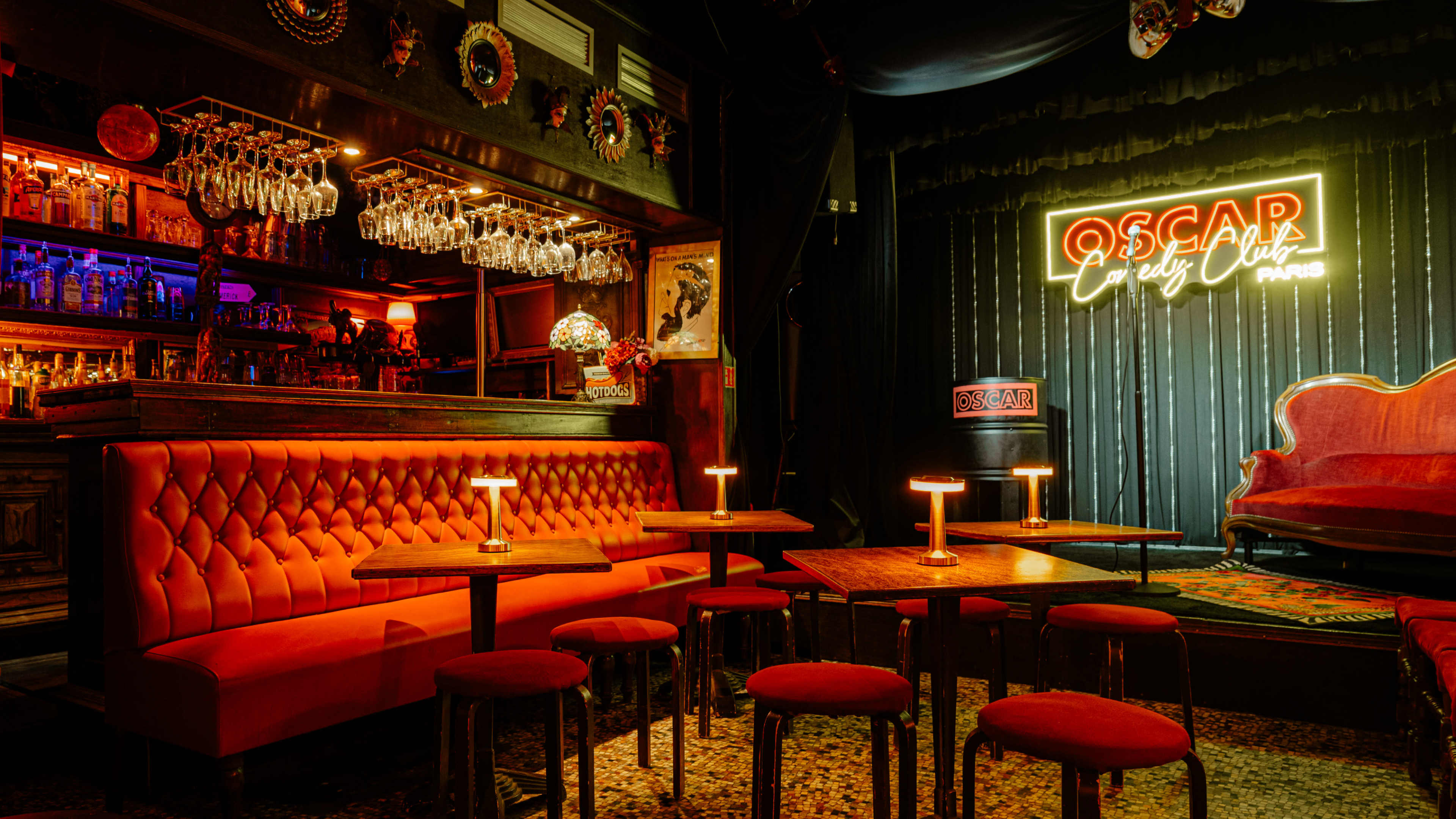 The image shows a bar area with red seating, small tables, and a neon sign that reads "OSCAR," set in a dimly lit, stylish interior.