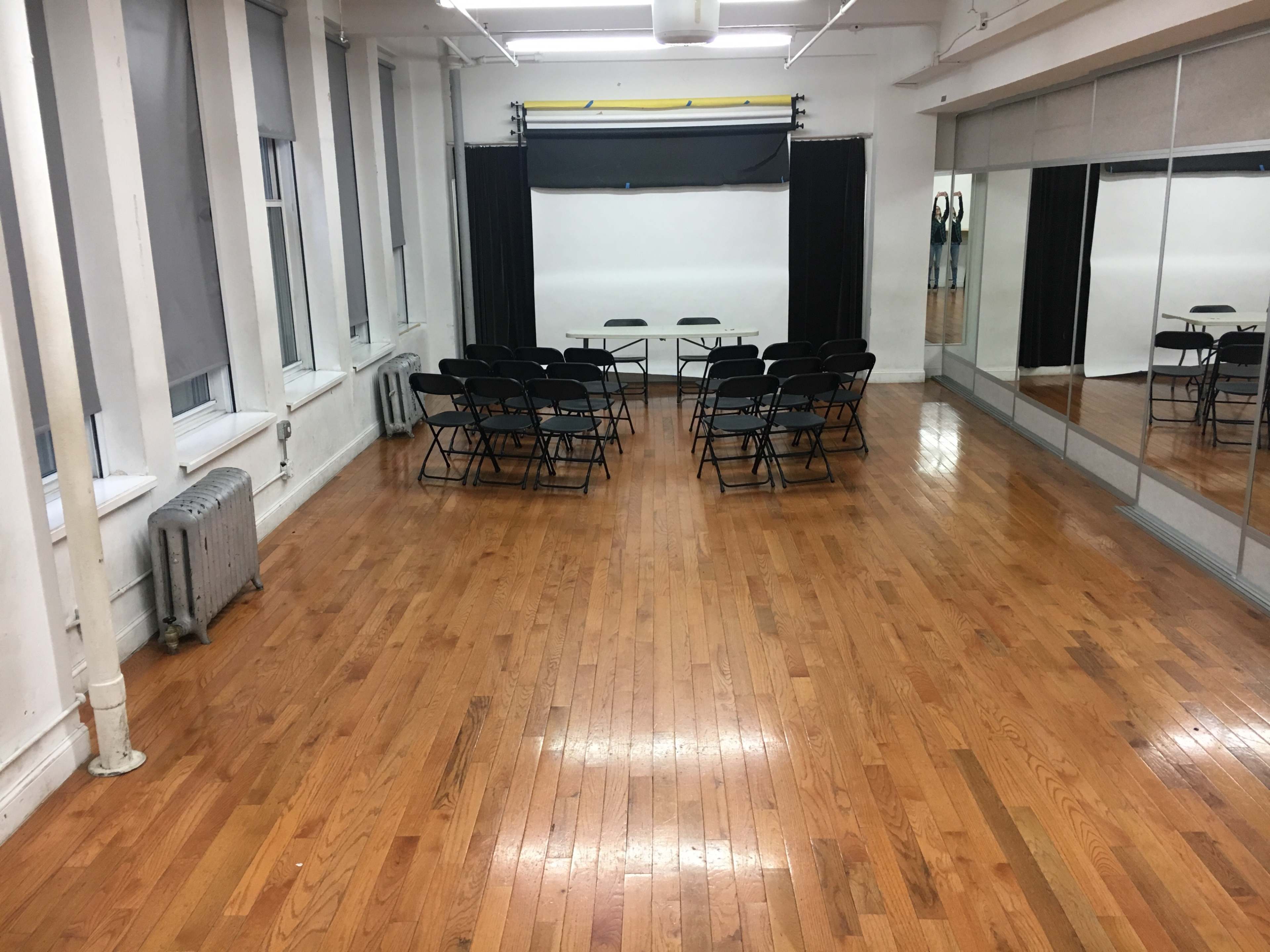 The image shows a spacious room with wooden floors, featuring several black folding chairs arranged in front of a table and a large blank screen on the wall.