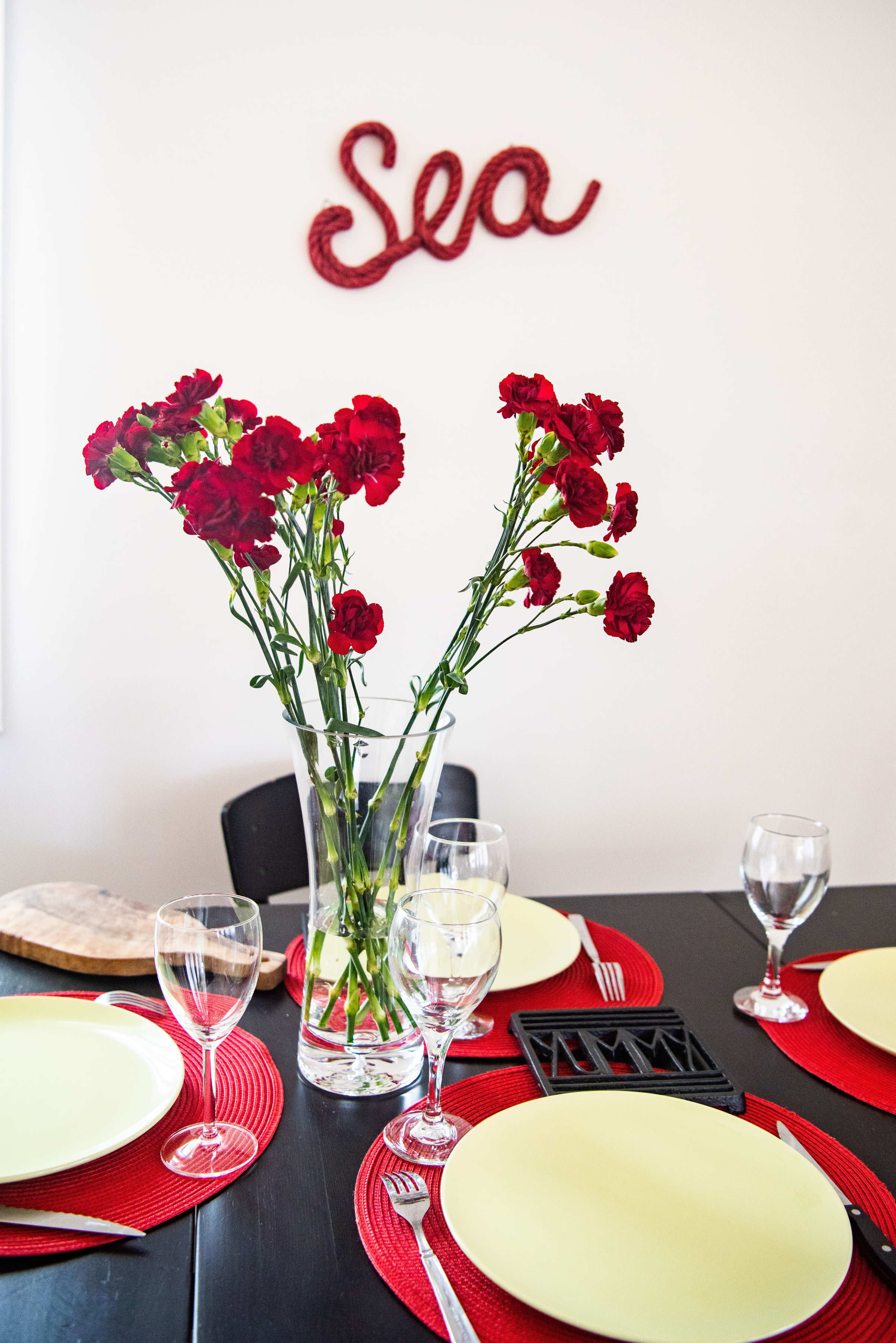 A dining table is set with yellow plates, glassware, and a vase of red flowers, while a decorative sign reading "Sea" hangs on the wall.
