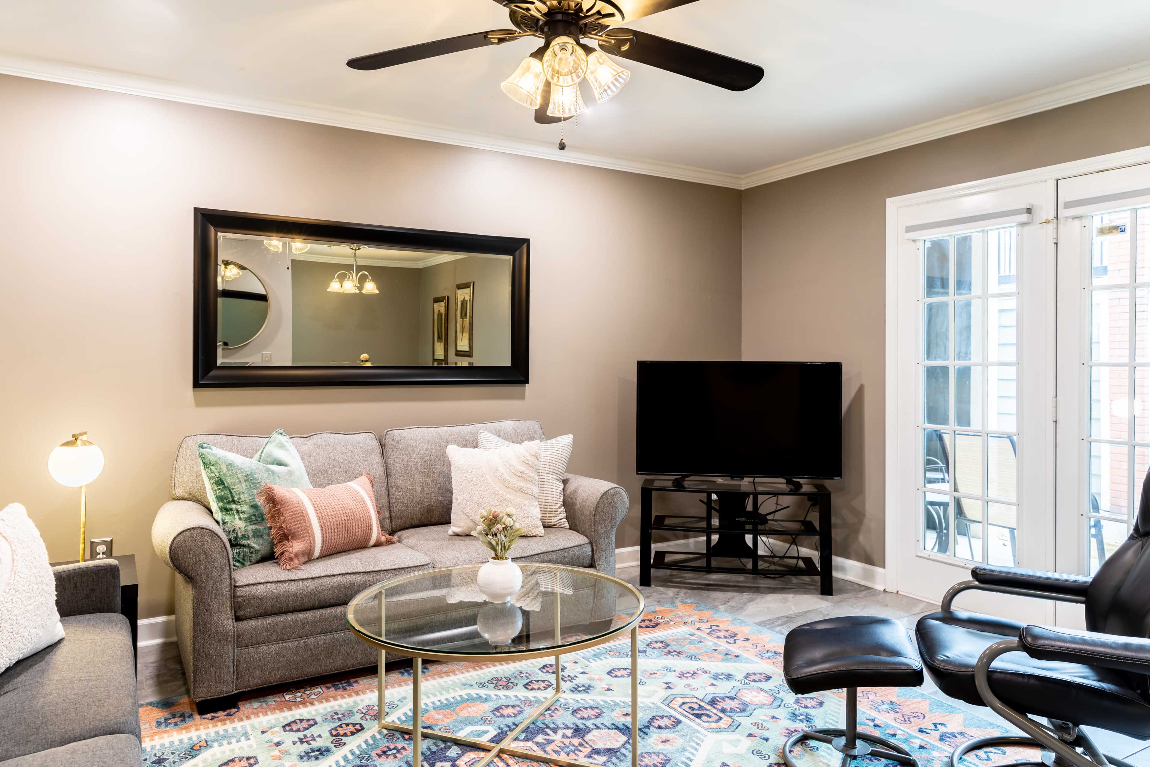 A living room features a gray sofa, a glass coffee table, a black television on a stand, and large windows with double doors leading outside.
