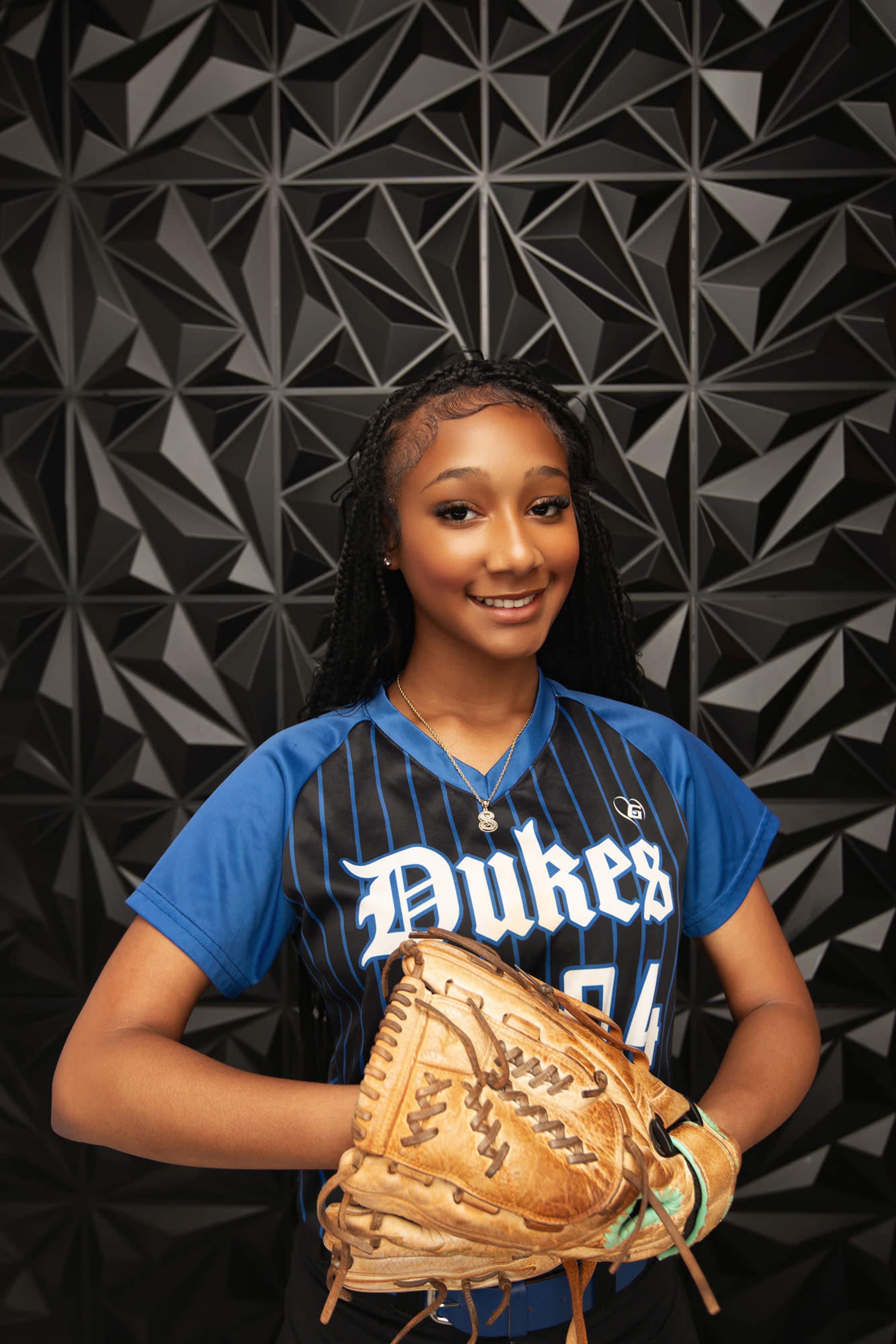 A young athlete wearing a blue and black softball jersey poses with a baseball glove against a geometric background.