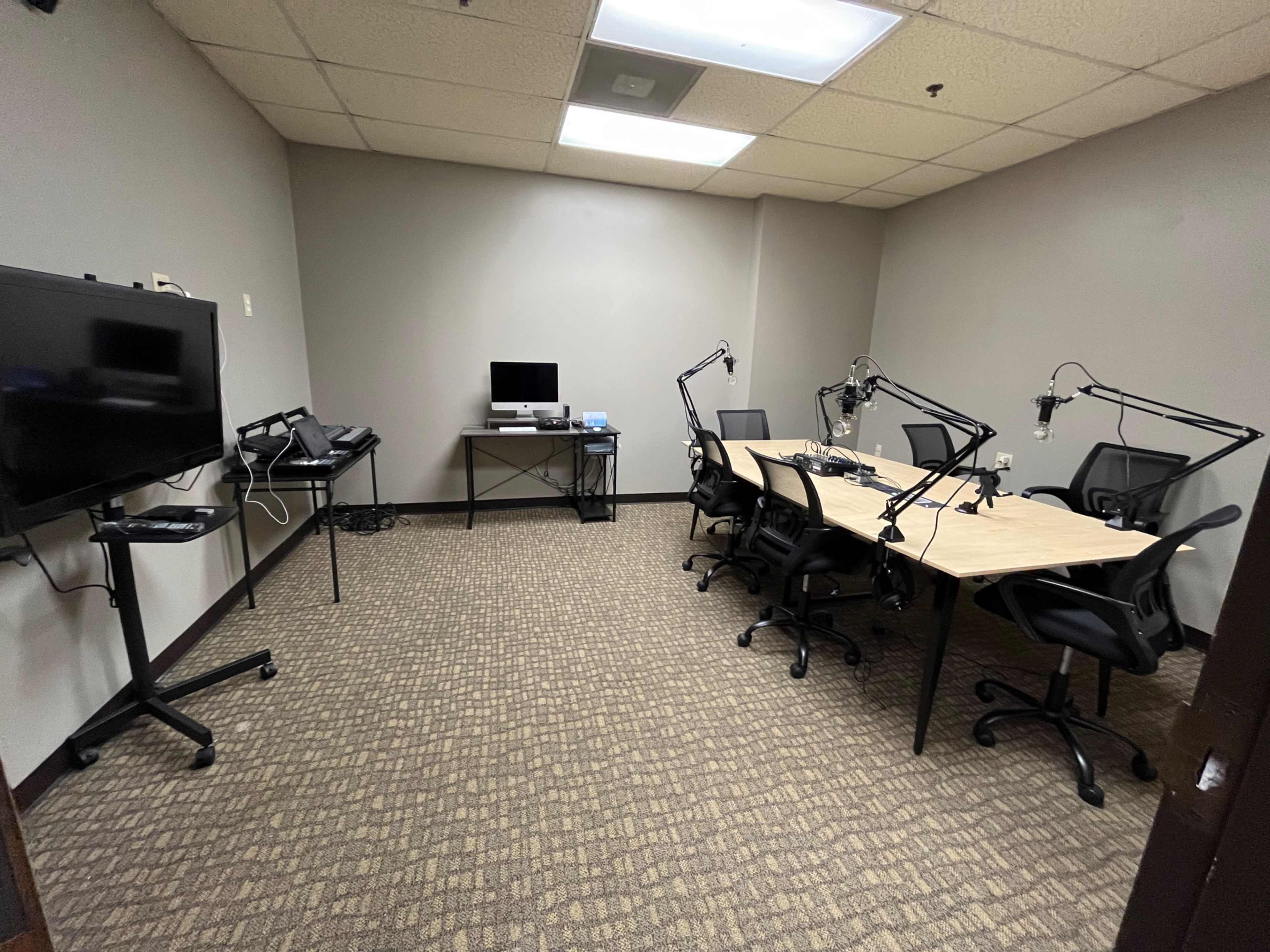 A small conference room features a row of desks with chairs, two monitors on stands, and several microphone arms attached to the tables.