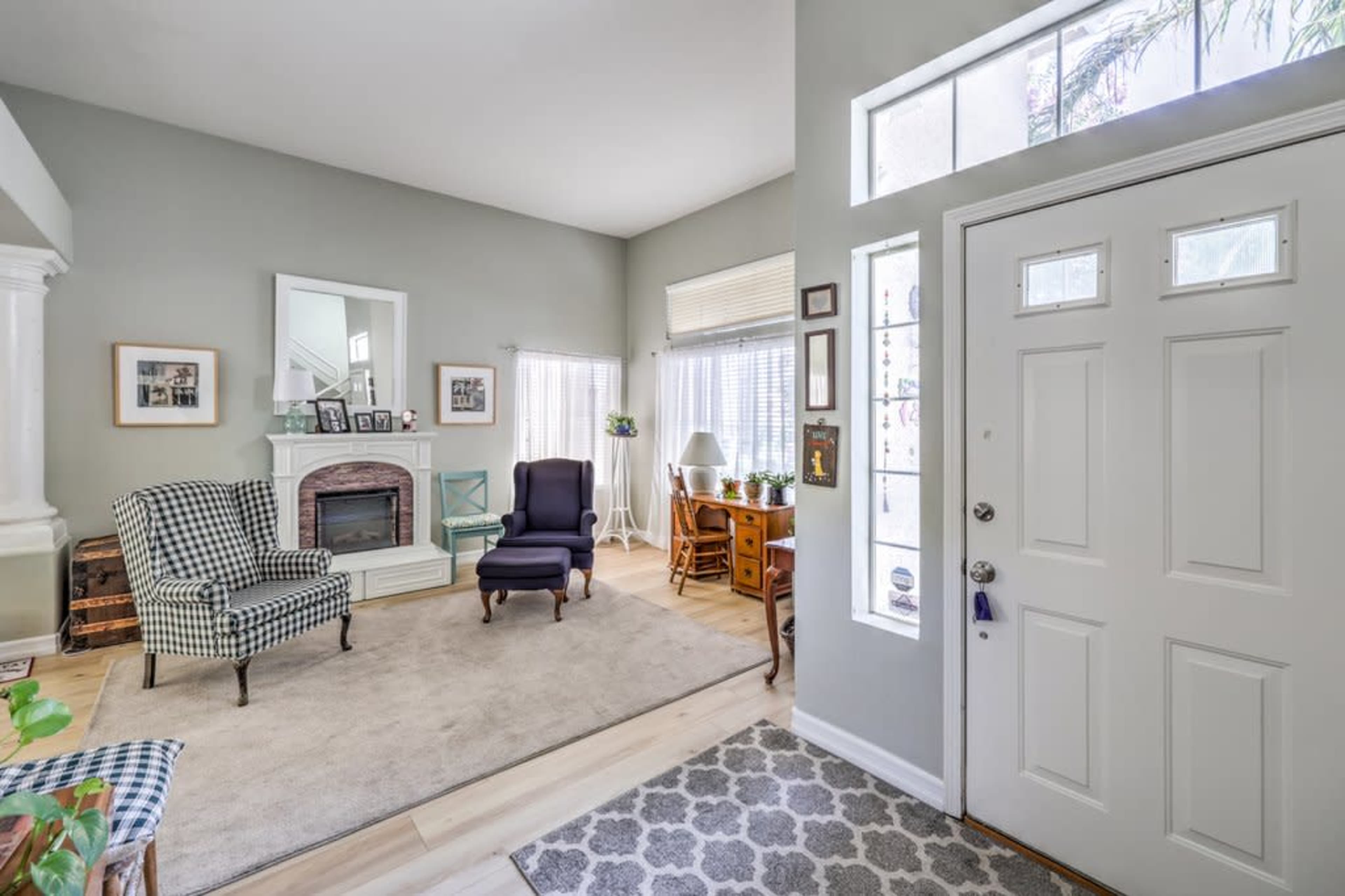 The living room features a fireplace, two armchairs, a small desk, and large windows that allow natural light to enter.
