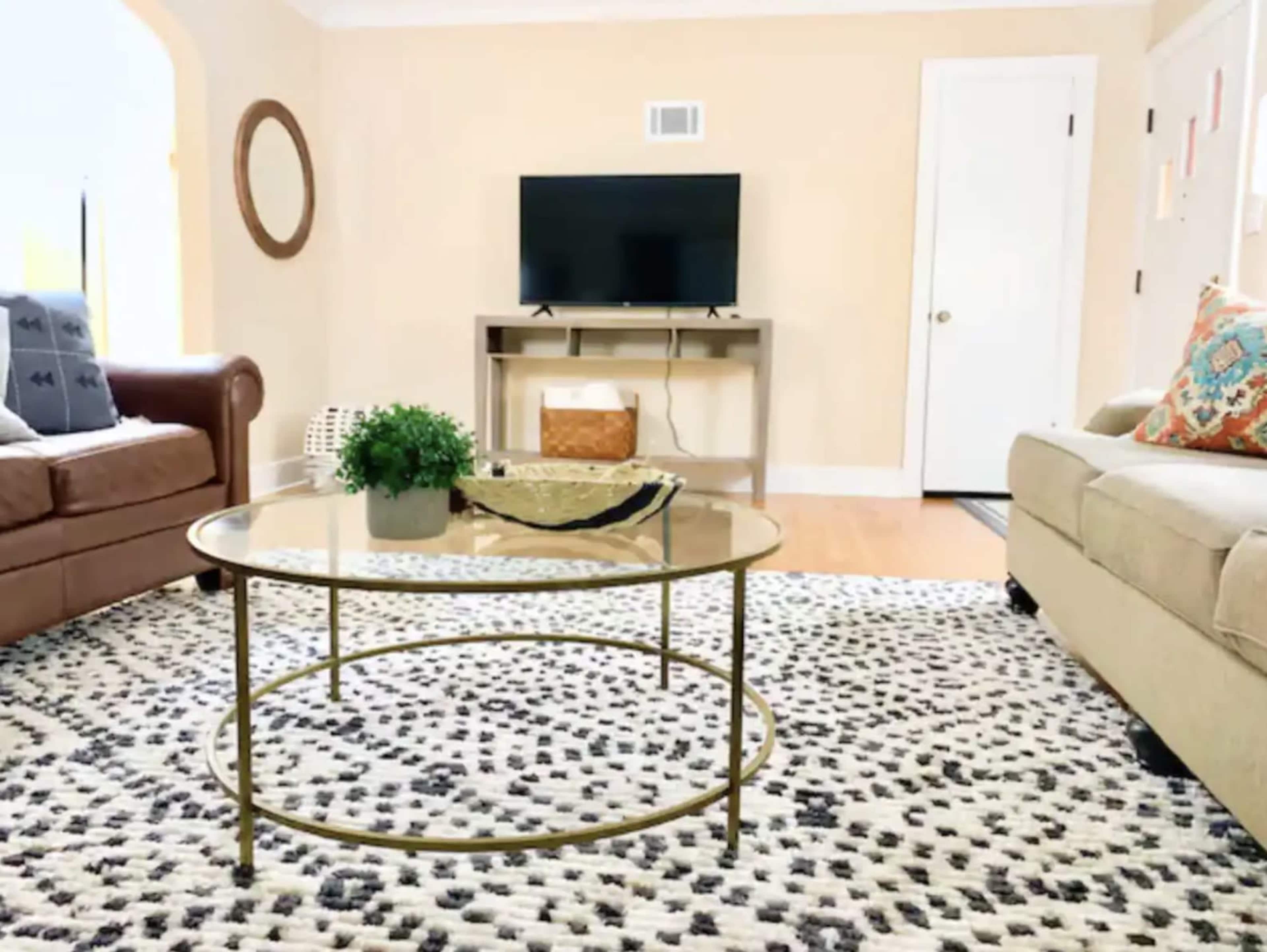 The living room features a glass coffee table on a patterned rug, flanked by a leather sofa and a beige couch, with a TV mounted on a stand against a wall.