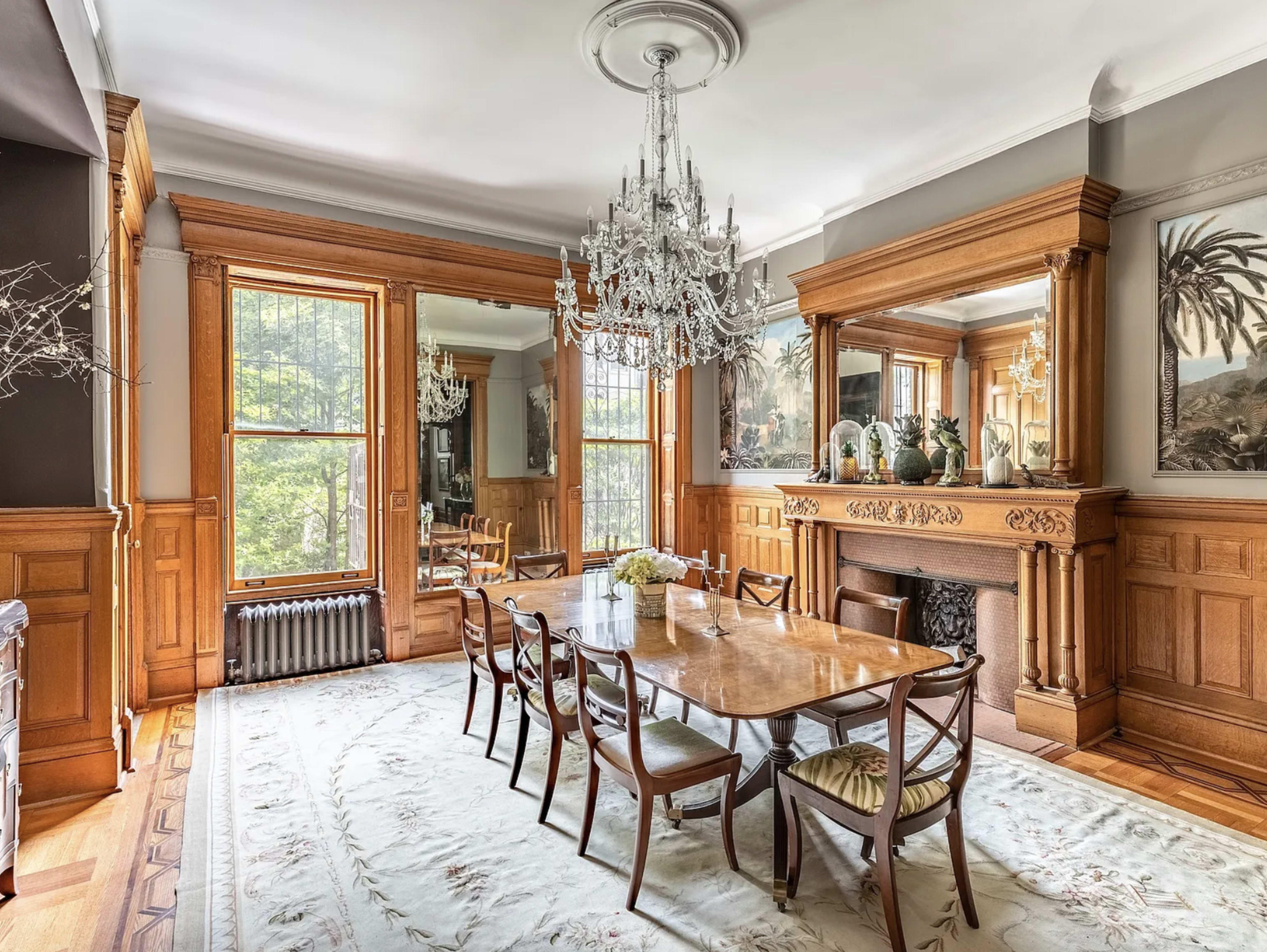 The image shows a dining room with a large wooden table surrounded by chairs, a chandelier hanging from the ceiling, and ornate woodwork on the walls and around the fireplace.