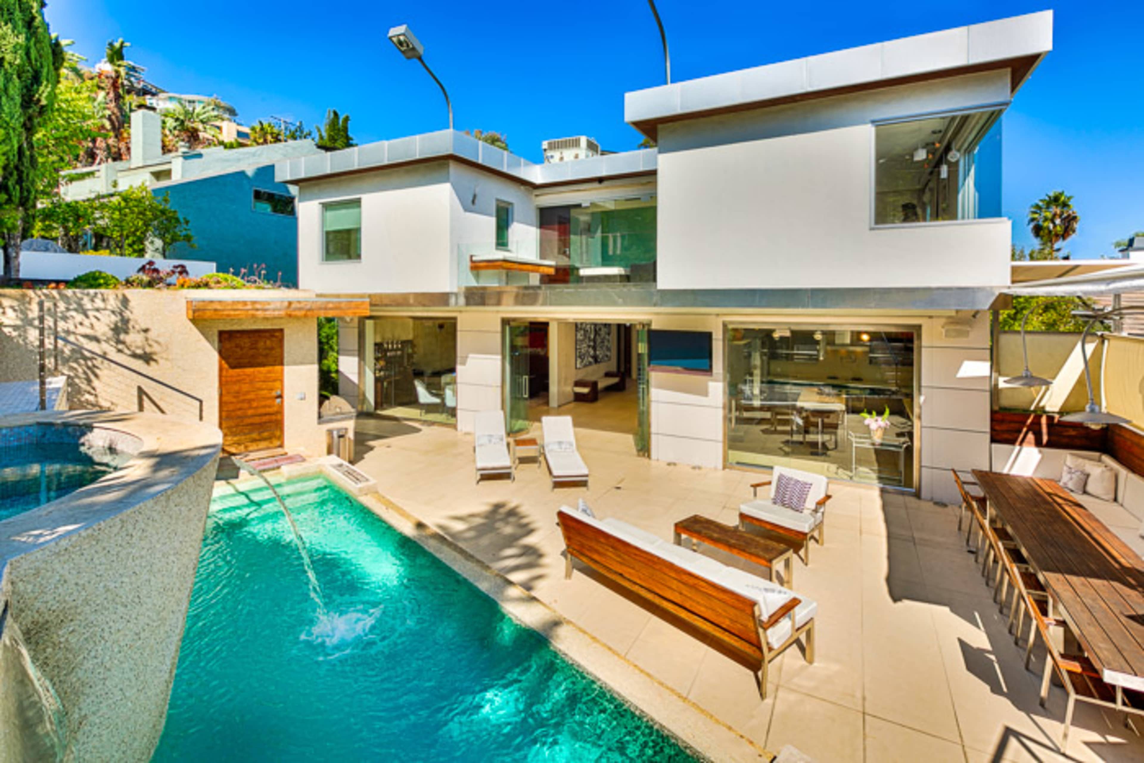 A modern two-story house features a pool, outdoor seating area, and large windows, surrounded by greenery.