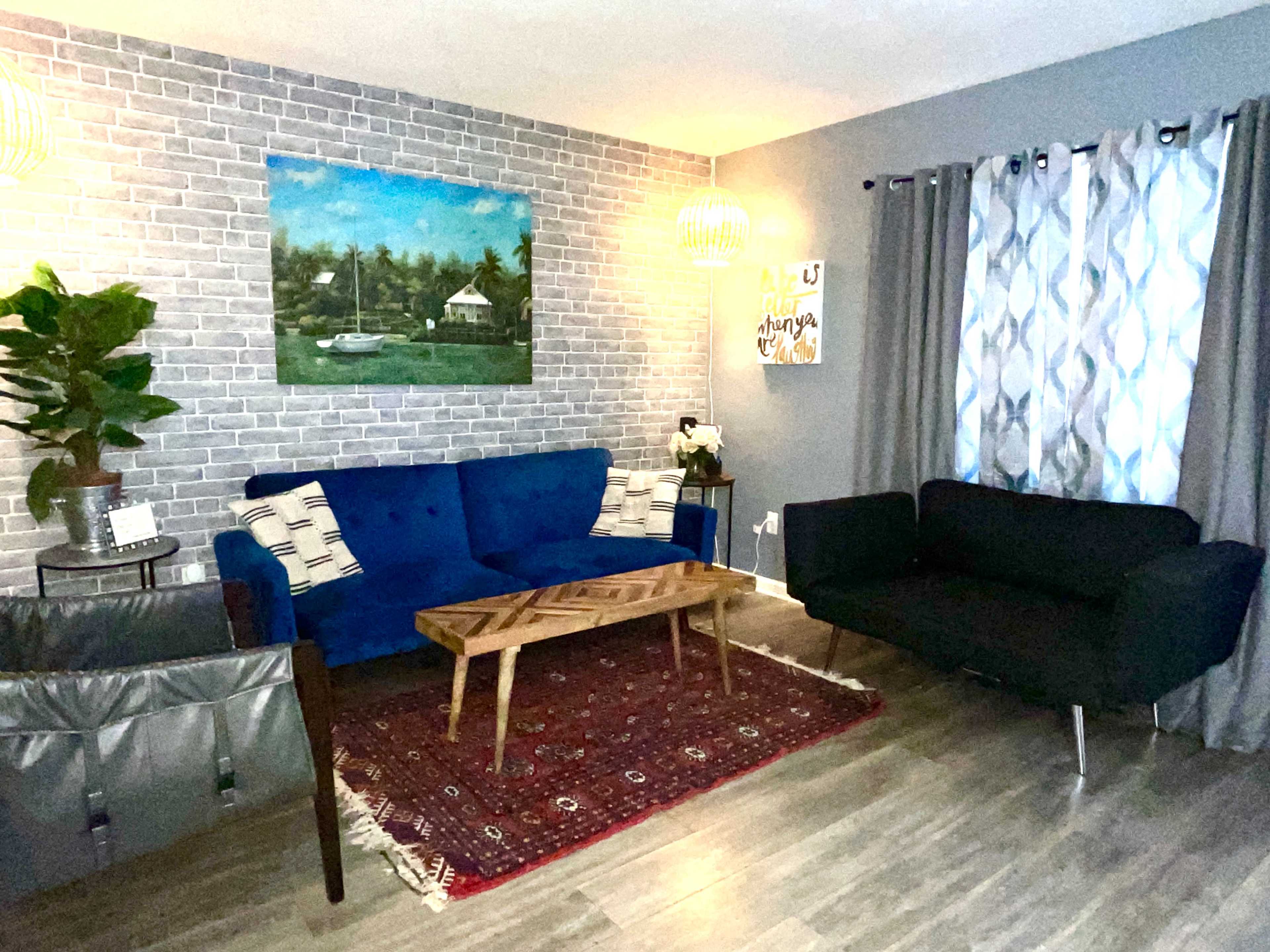 The image shows a living room with a blue sofa, a black loveseat, a wooden coffee table, and a large wall painting, all arranged on a hardwood floor with an area rug.
