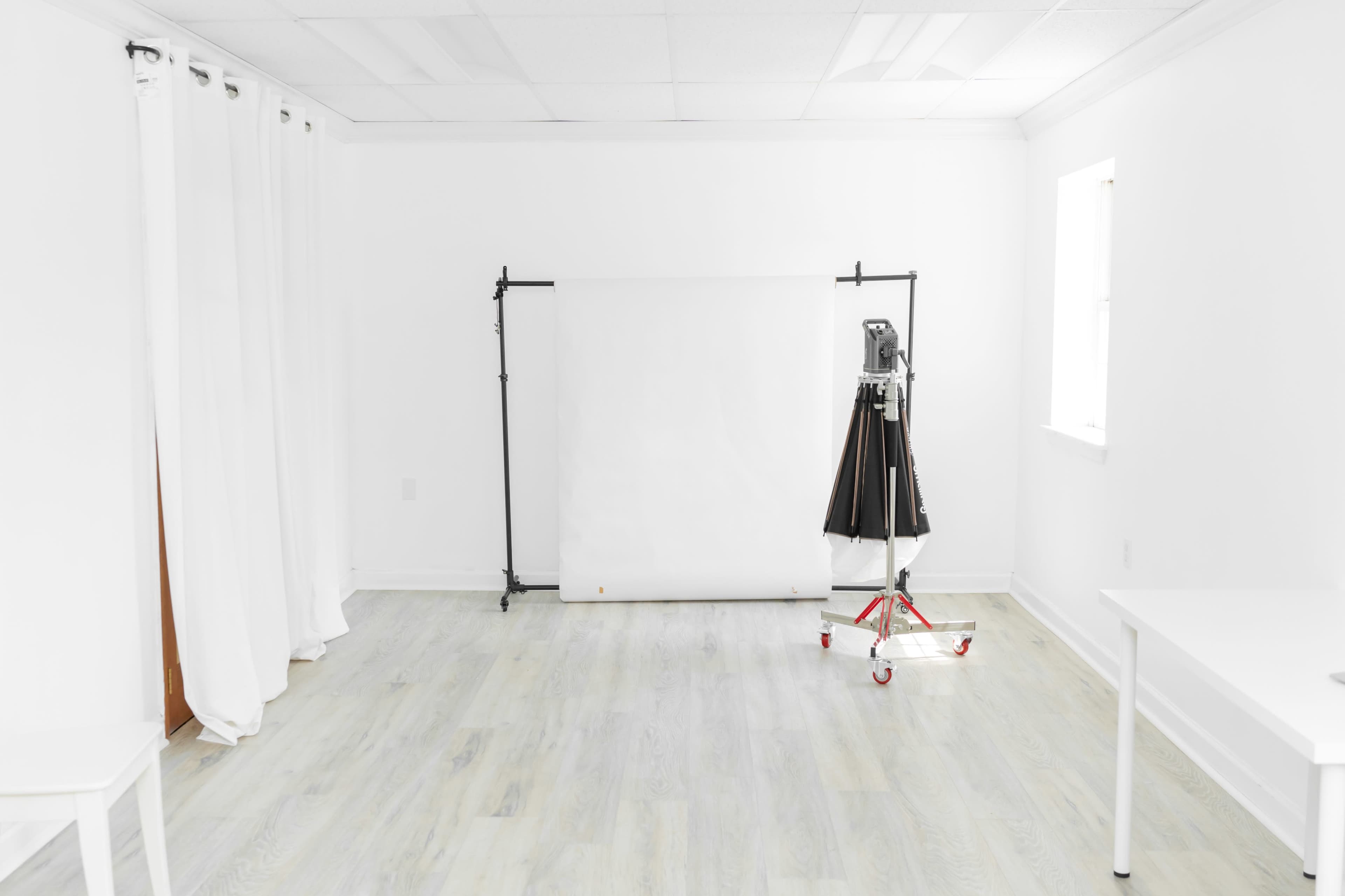 A bright, minimalist studio features a blank white backdrop, a lighting rig on wheels, and white curtains along one wall.