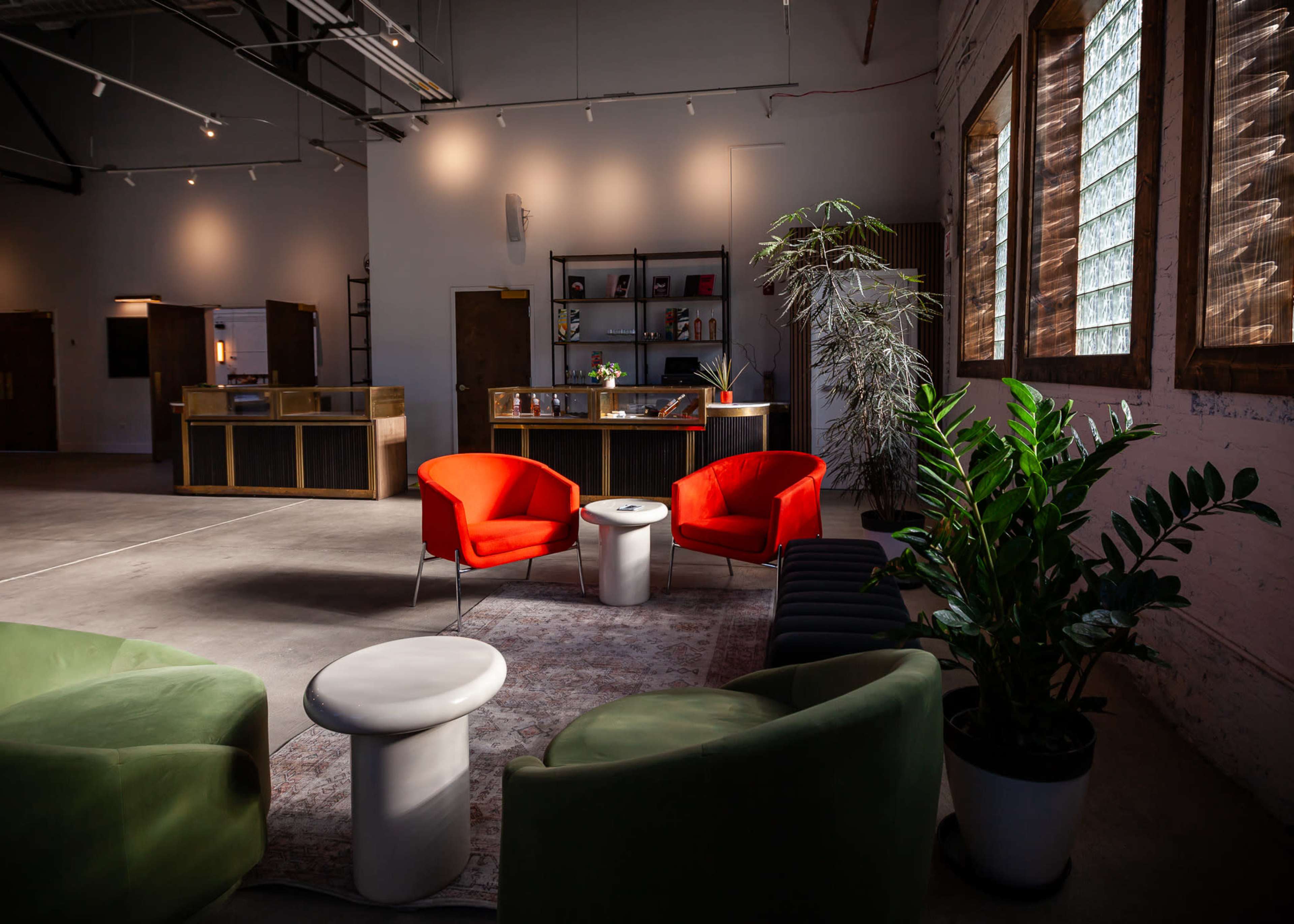 The image shows a modern lounge area with orange and green chairs, a circular white table, and large windows allowing natural light to enter the space.