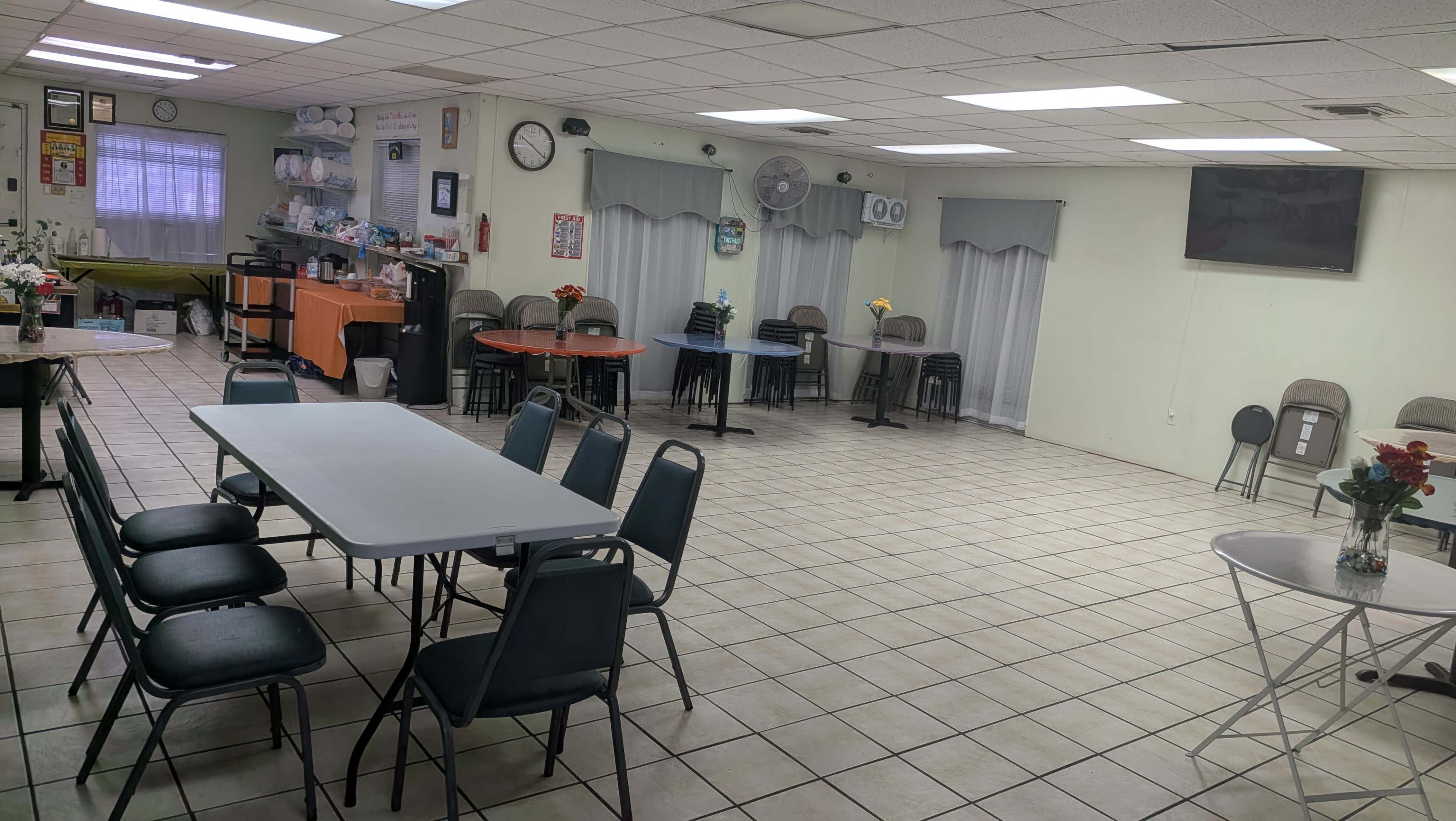 A spacious, well-lit room features several tables and chairs arranged throughout, with a TV mounted on the wall and a small refreshment station in the corner.