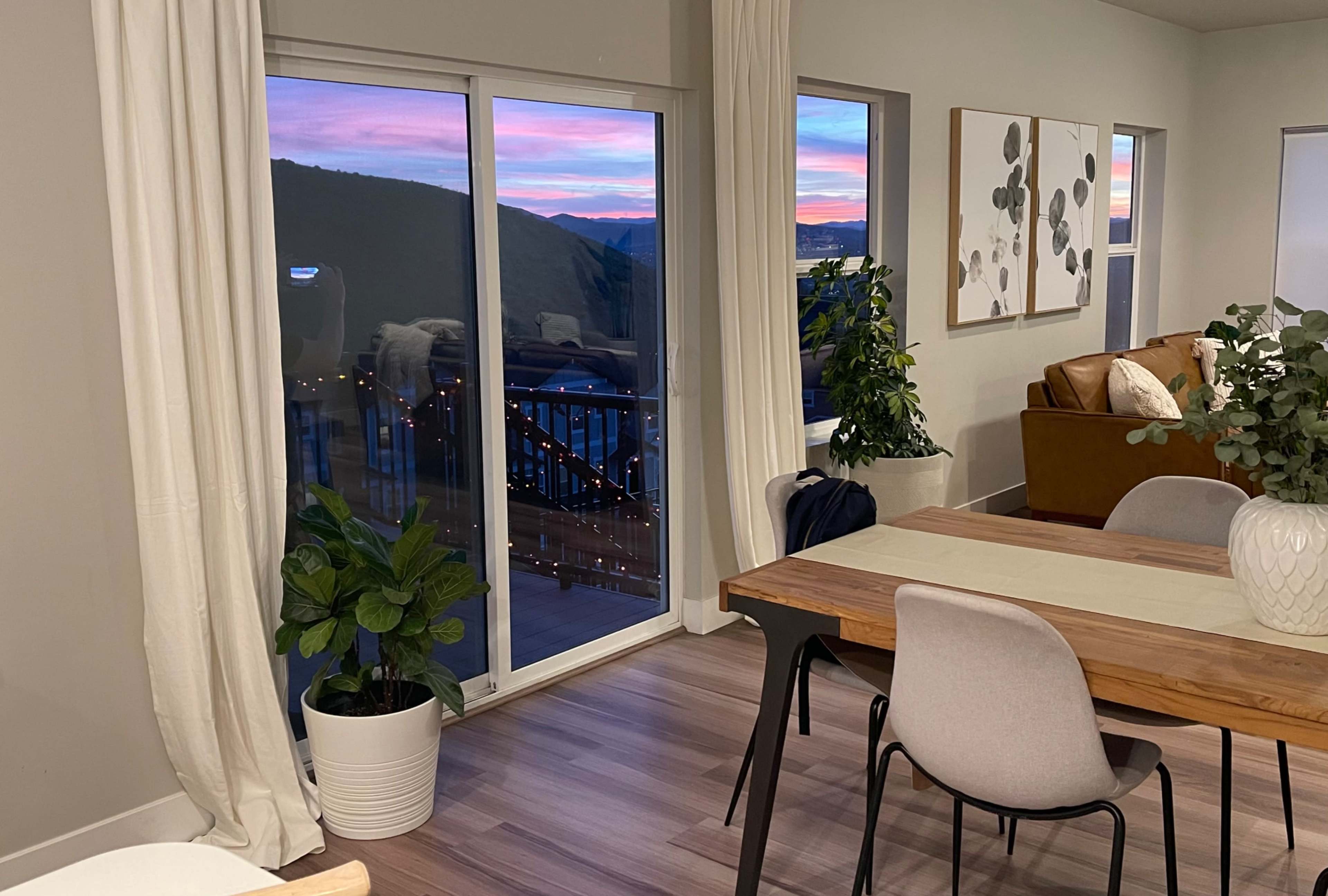 A modern living space with a dining table, plants, and large windows revealing a sunset view outside.
