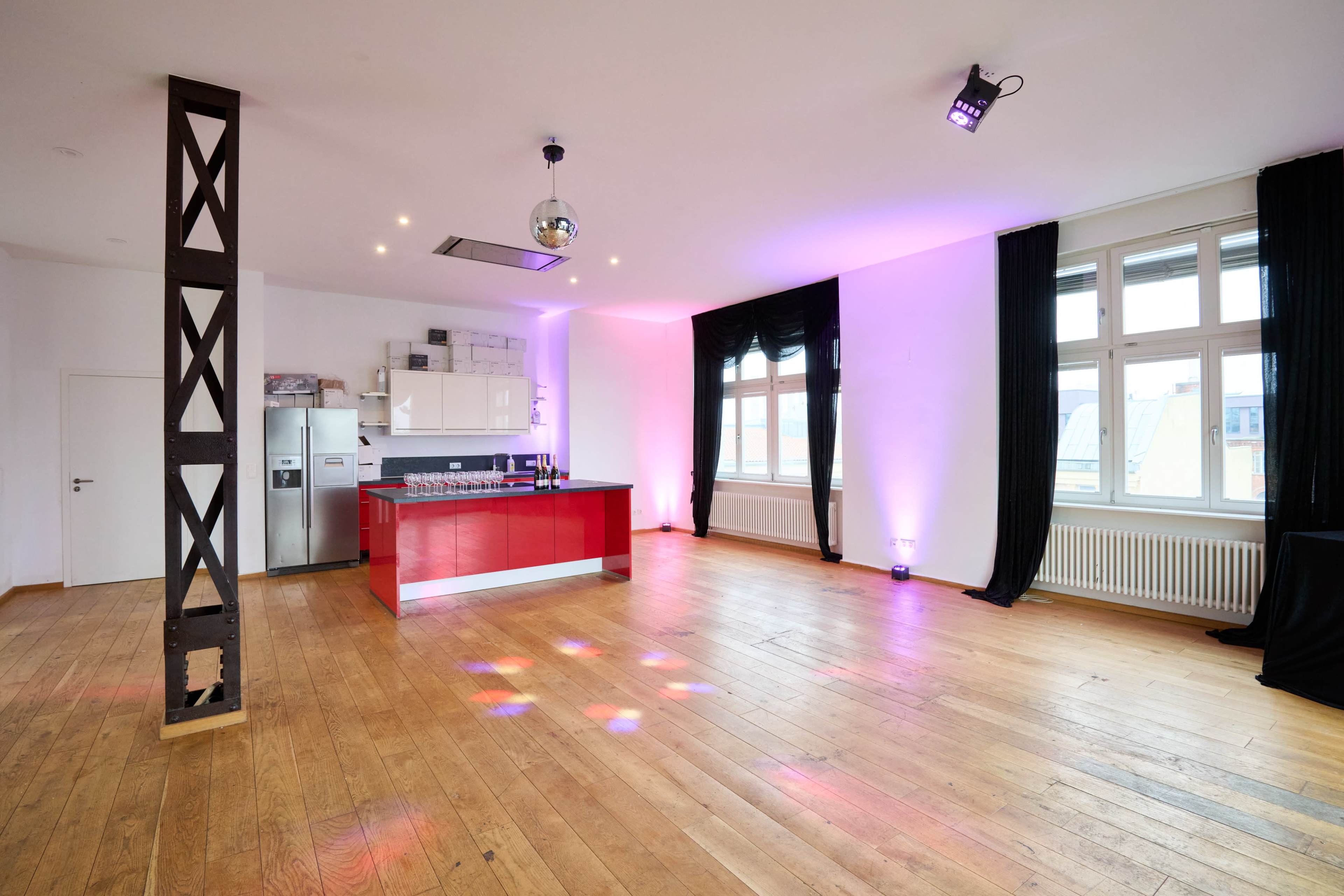 The image shows an empty room with a wooden floor, featuring a kitchen area with red cabinetry and stainless steel appliances, illuminated by colored lights.
