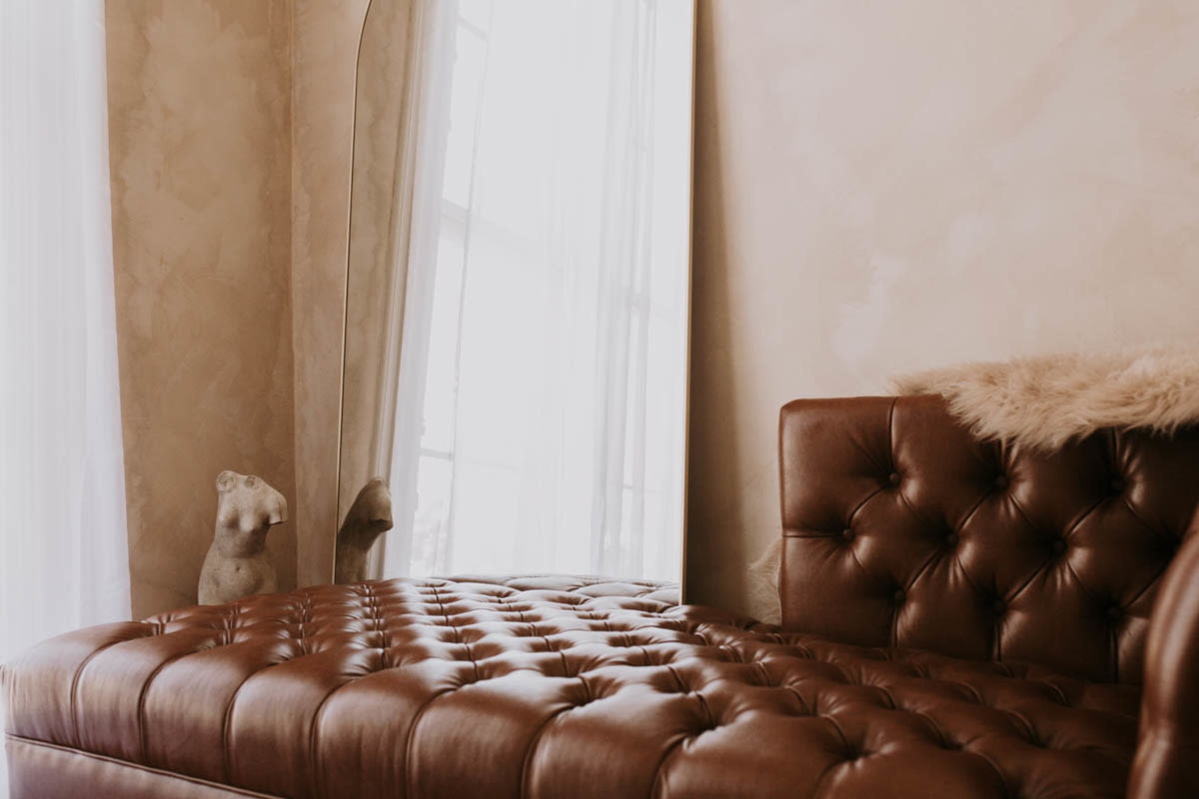 A tufted brown leather chaise lounge is positioned beside a window with sheer curtains, while a sculpture of a figure is visible in the background.