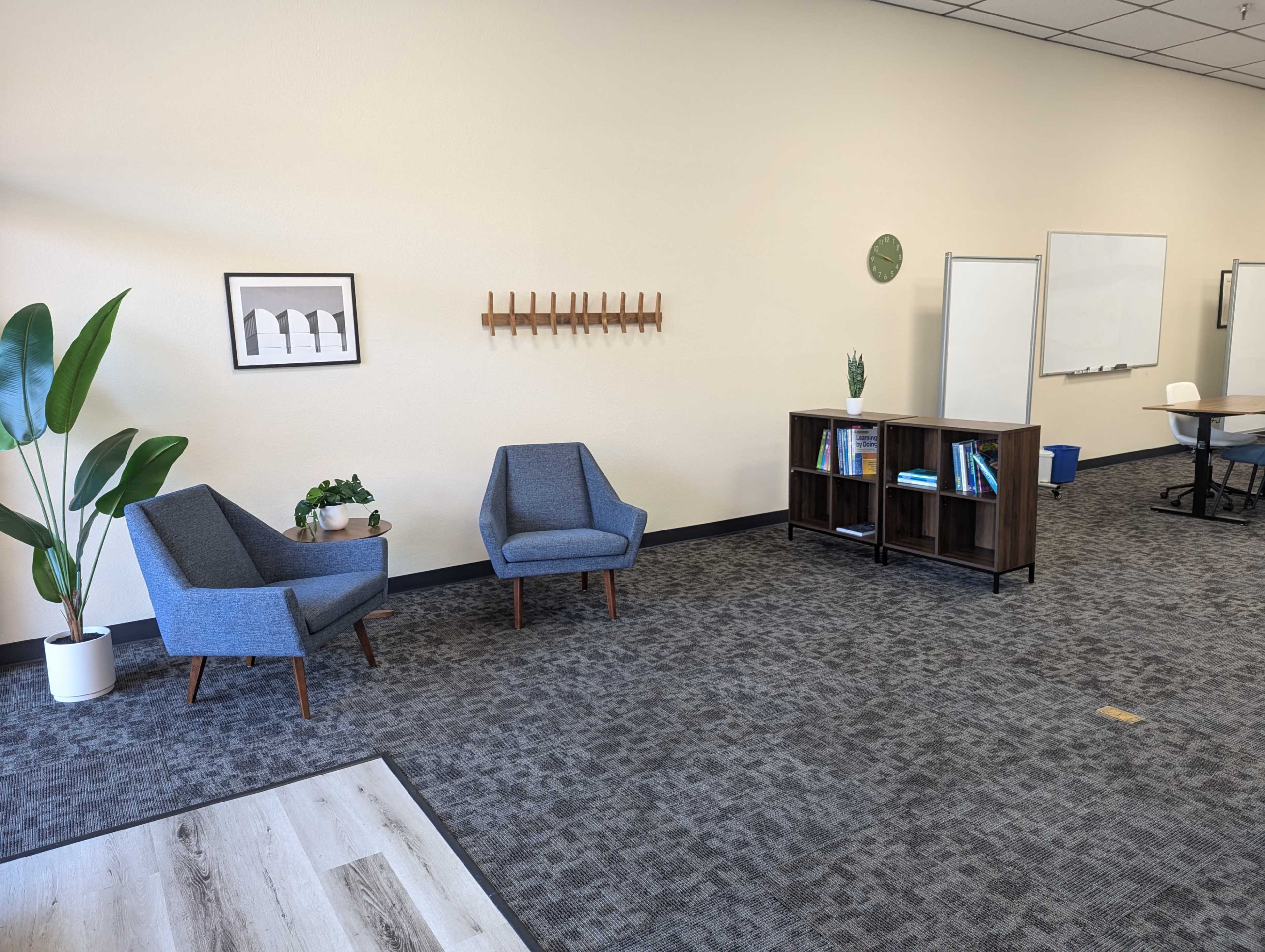 A modern office interior featuring two blue chairs, a wooden bookshelf with books, and a whiteboard against a plain wall.