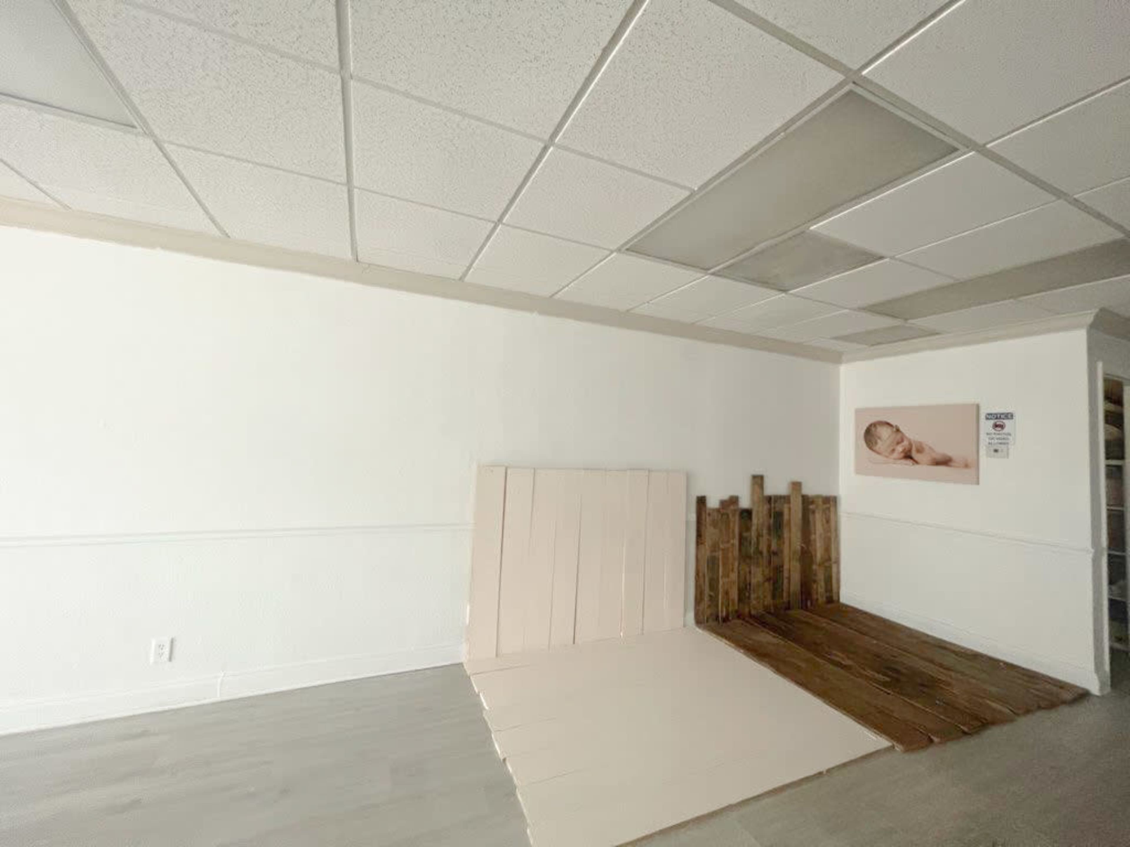 The image shows an empty room with a textured white wall, a section of light-colored wooden flooring, and a wall display featuring a photograph.