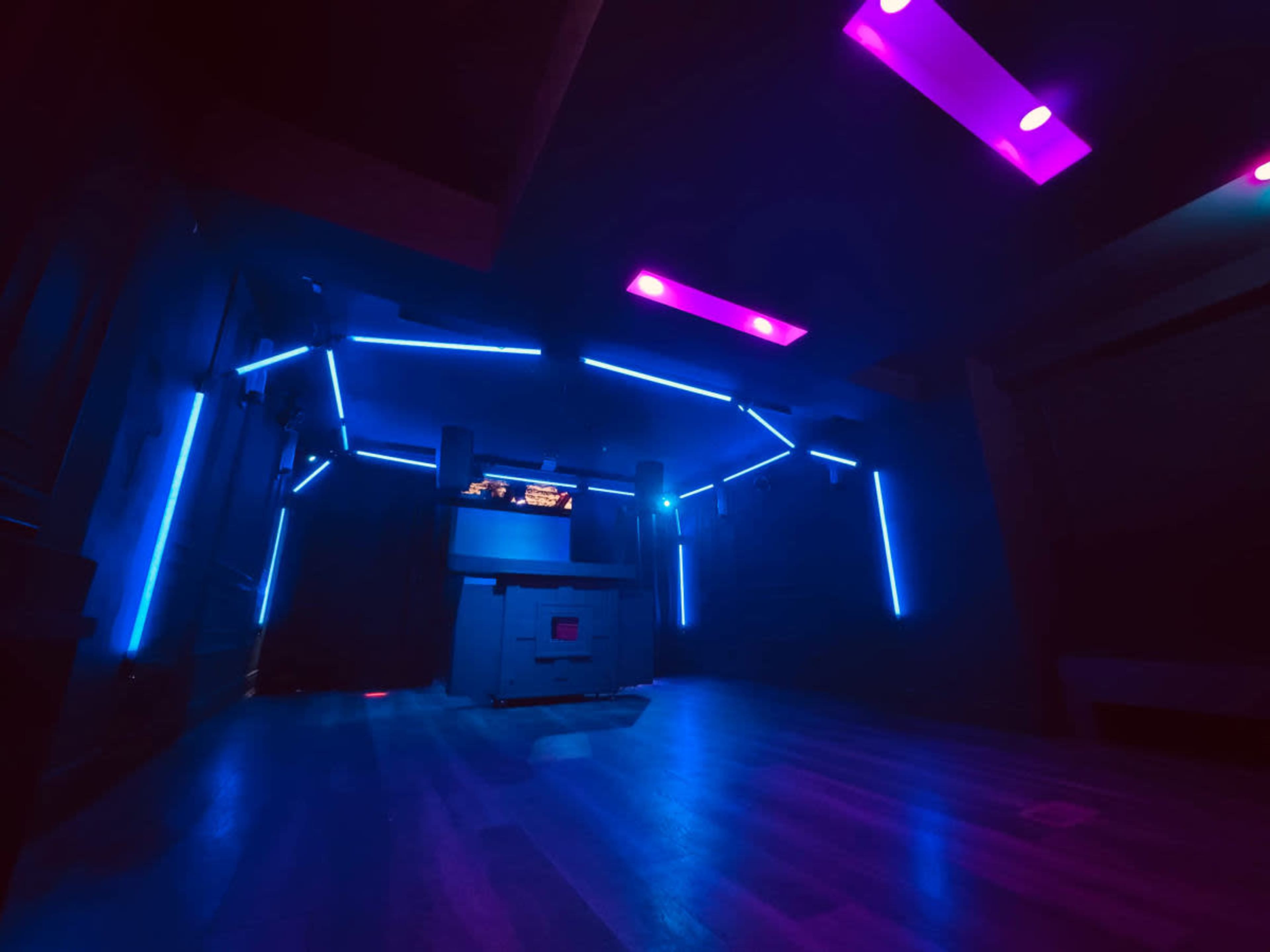 The image shows a dimly lit room illuminated by blue and pink LED lights with a central structure that appears to be a DJ booth or sound system.