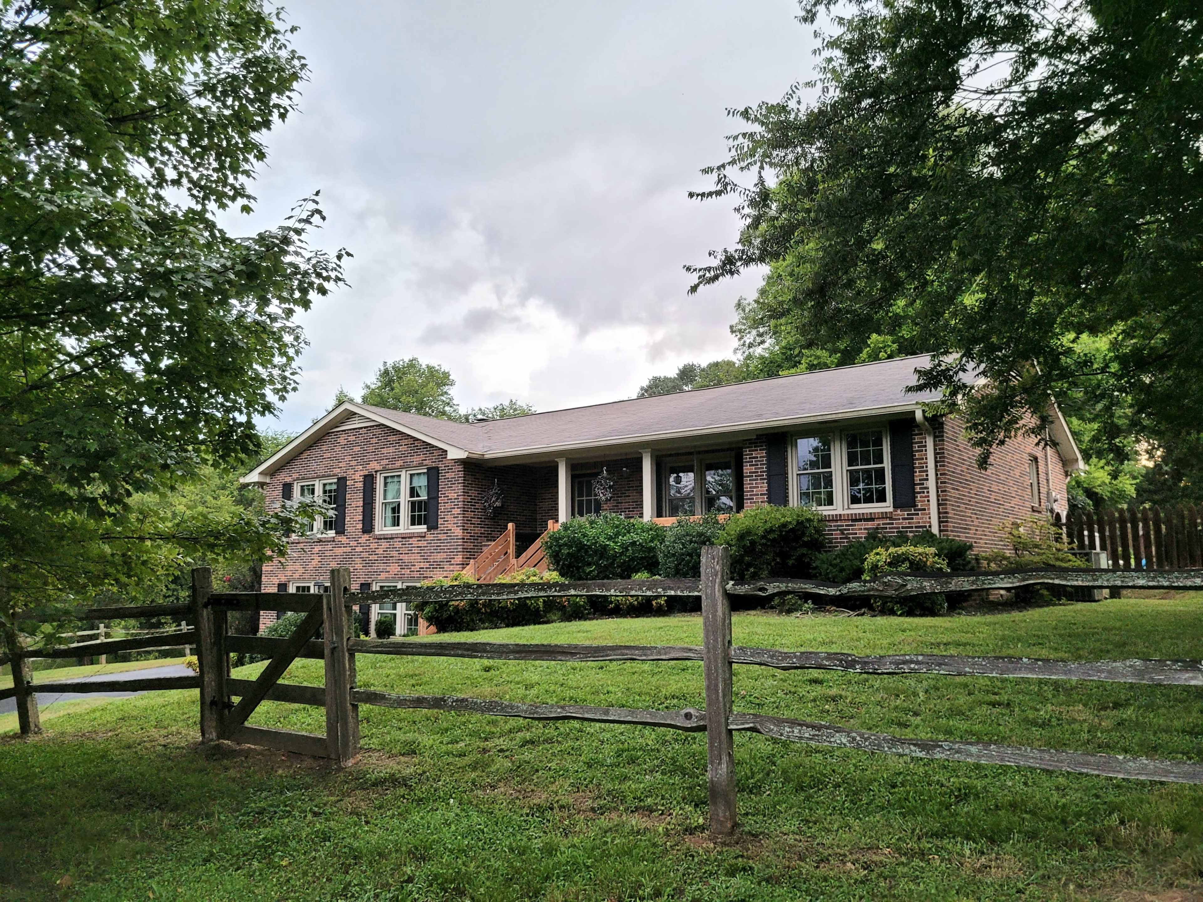 A brick ranch-style house sits behind a wooden fence, surrounded by greenery under a cloudy sky.
